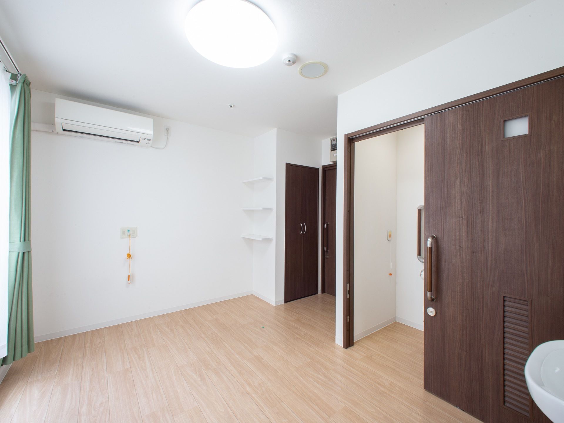 右奥にトイレがあり、その隣に入口扉と収納スペースがある。壁には３段の棚が取り付けられている。天井近くにエアコンがある。