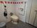 白色の手すりが両脇に設けられた、洋式トイレである。背もたれ用のクッションも備え付けられている。ゴミ箱もある。