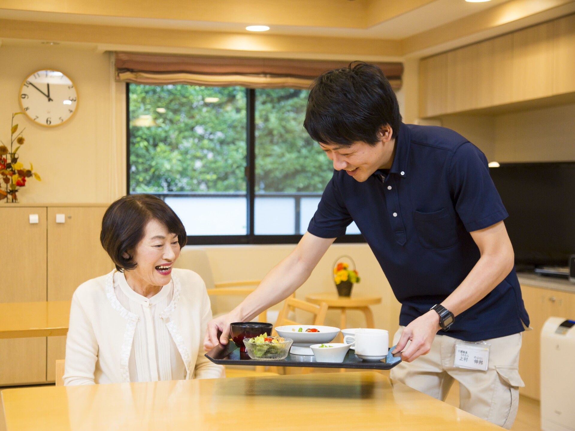 テーブルに座り待っている女性入居者に食事を運んでいる男性スタッフ。食事内容を見て笑顔になる女性。
