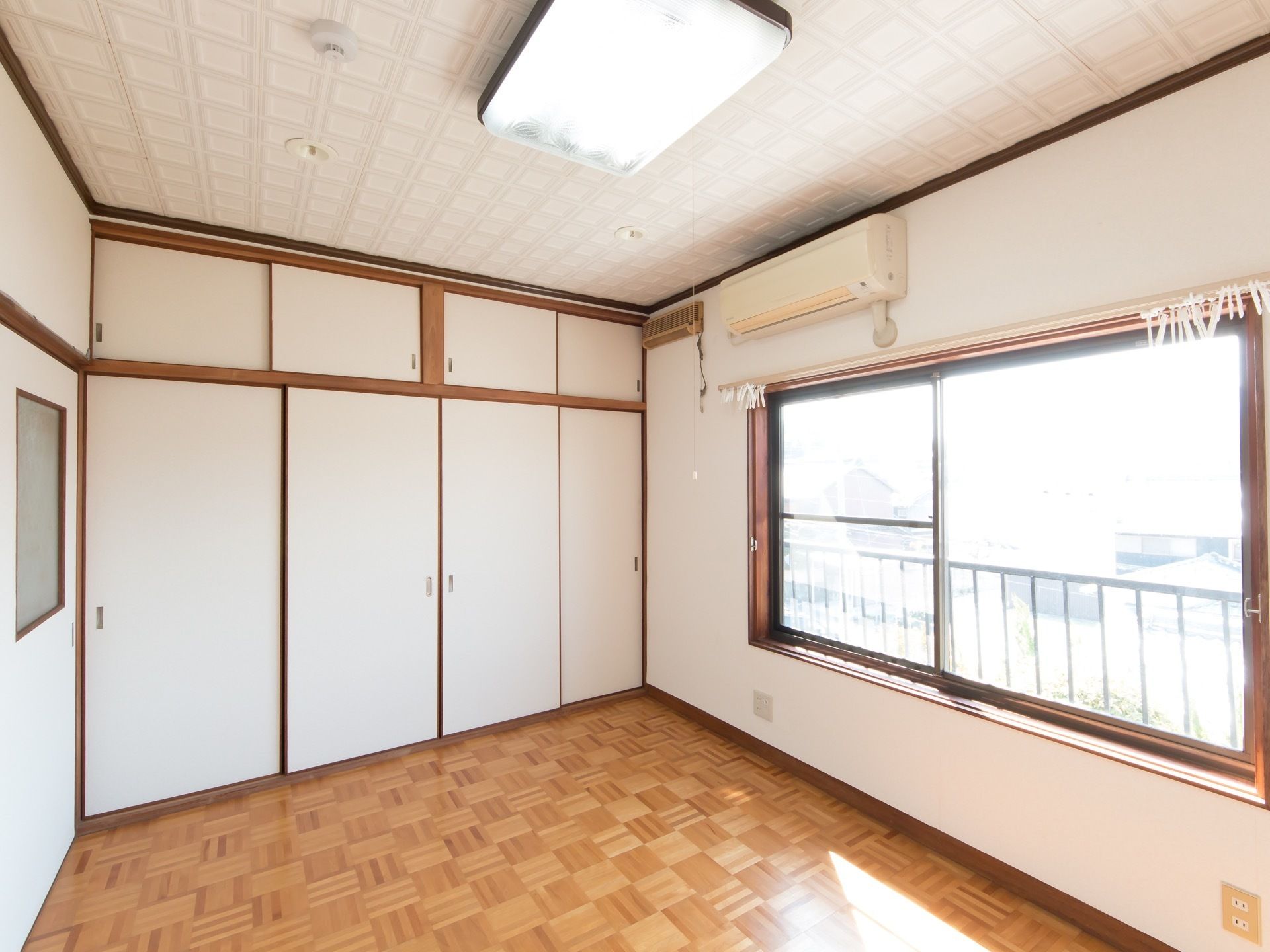 四角い木目のタイルが交差するようなデザインで敷かれている洋間がある。右手には窓があり、窓の上にエアコンが設置されている。