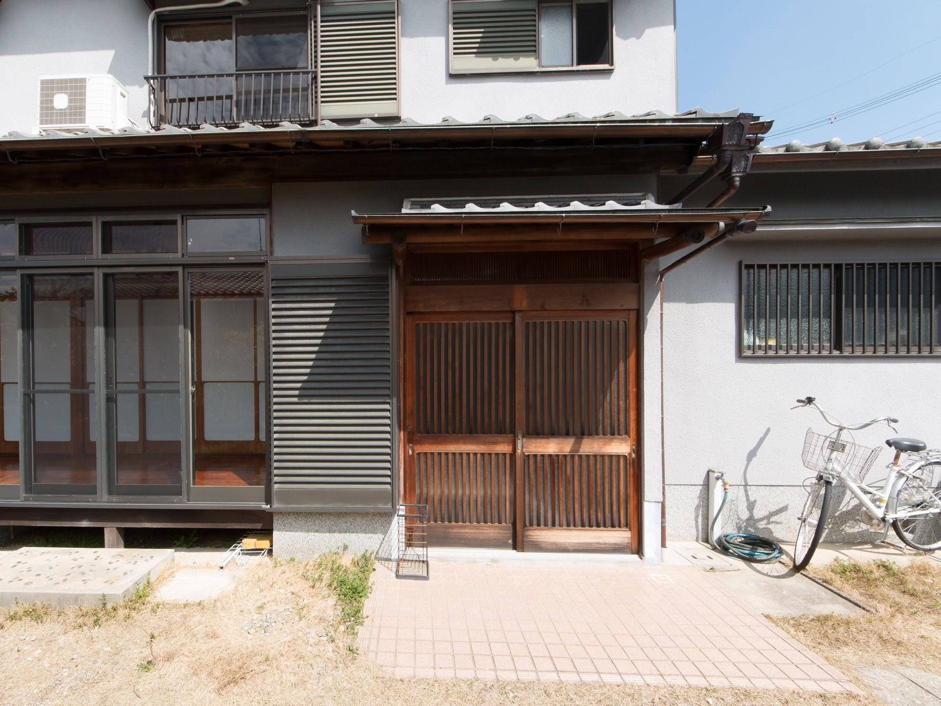 木製の格子扉の引き戸の入口で、瓦屋根がせり出している。入口前に傘立てが置かれ、右手に自転車が停まっている。