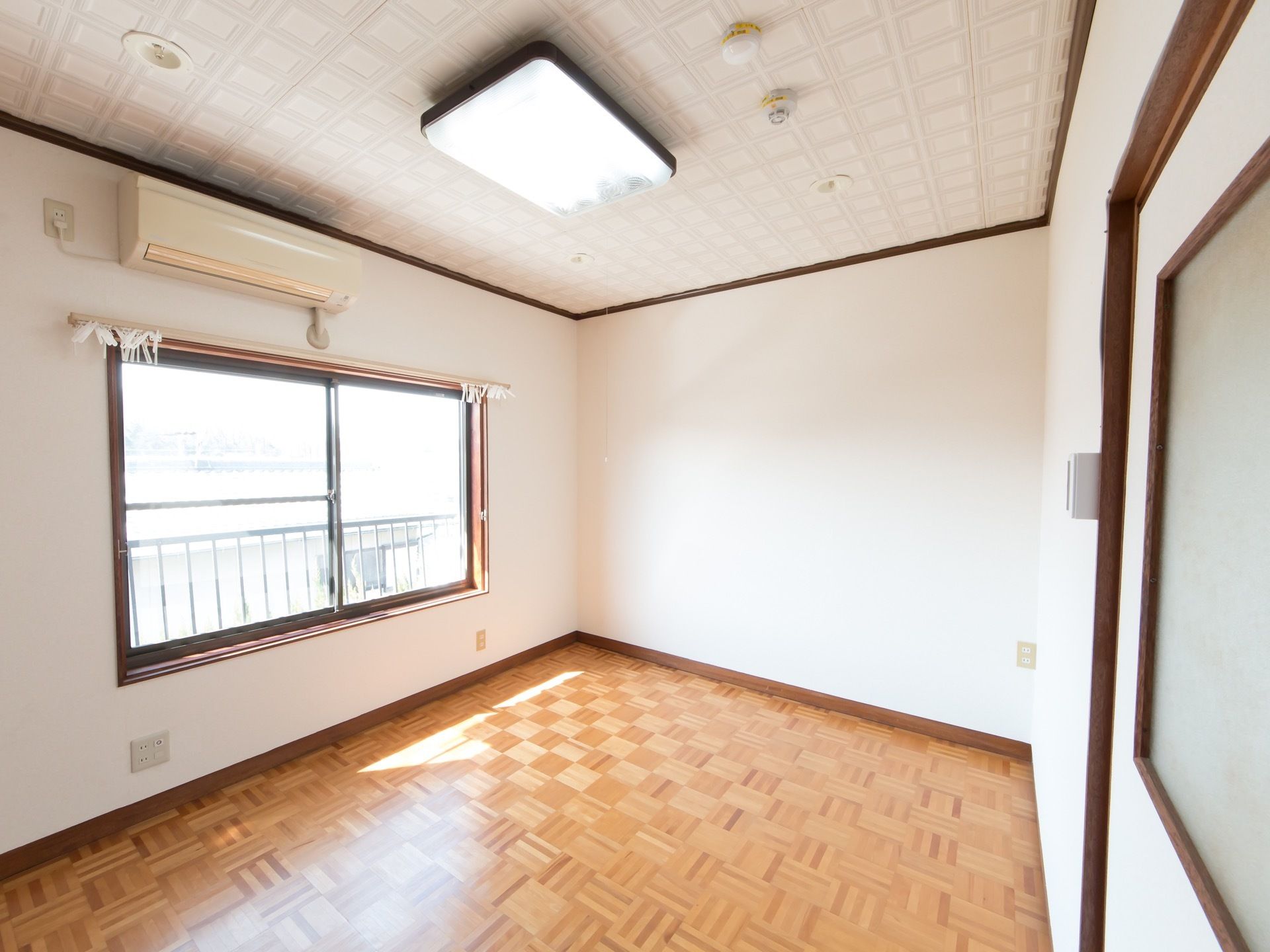 白い壁で囲まれた居室で、左側に大きな窓が開いている。窓の外には落下防止の柵がある。窓の上にエアコンがある。