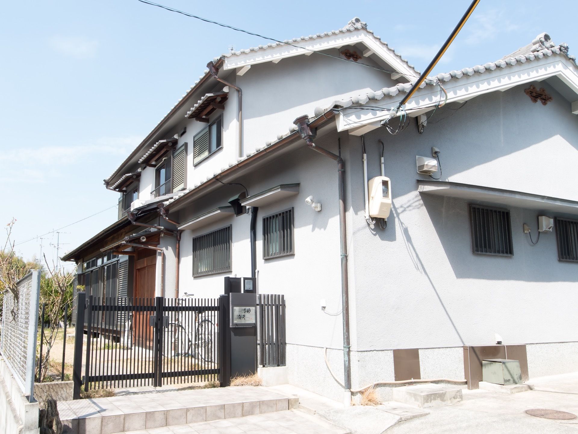 白い外壁の２階建ての建物で、瓦屋根が乗っている。左手前に黒い門扉が閉まっており、施設前は駐車スペースがある。