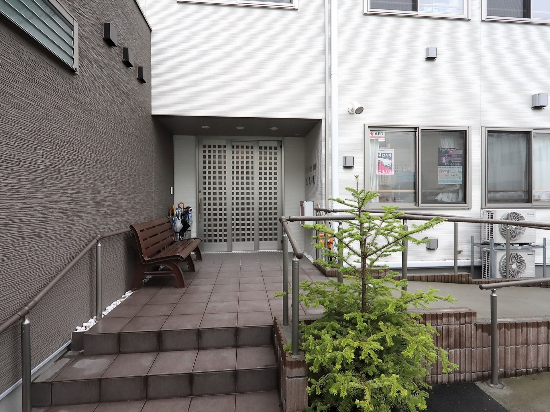 エントランスには、手すりが備わった階段とスロープがある。ベンチと傘立てが置かれており、壁に郵便受けが設置されている。