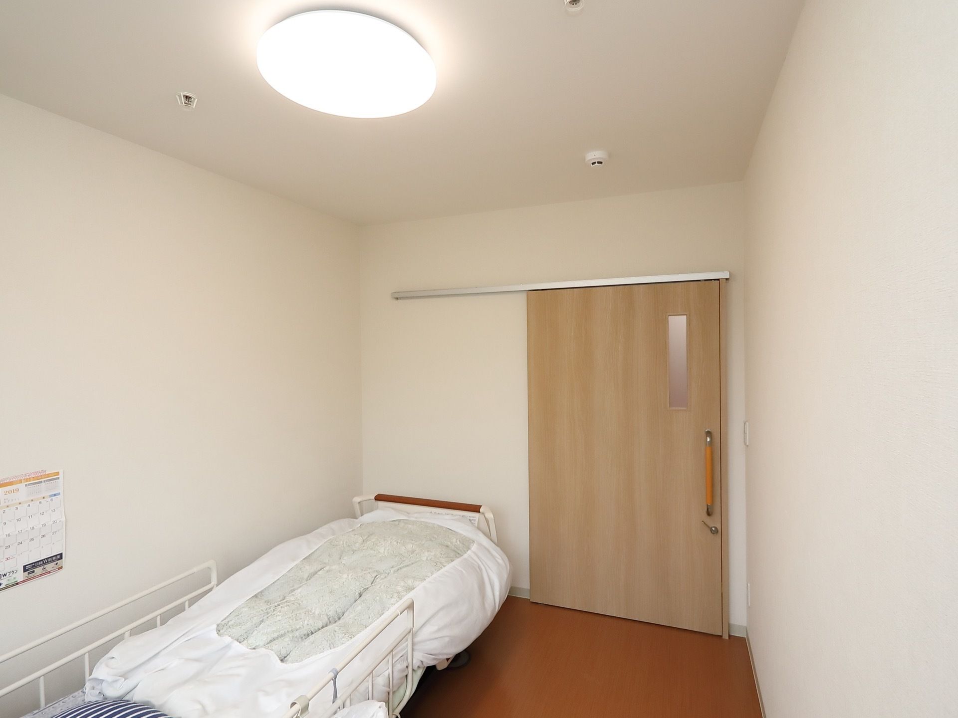 個室の居室である。介護ベッドや照明、スプリンクラーが完備されている。出入口の扉は引き戸になっている。