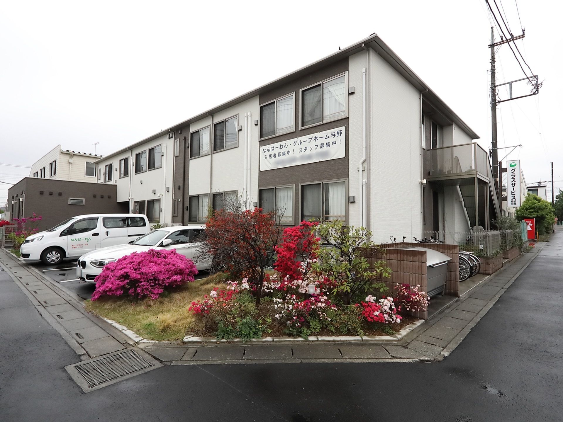 白とグレーを用いた外装のホームの前面に、駐車場を完備している。駐車場の脇の植え込みに、季節の花が咲く庭木がある。
