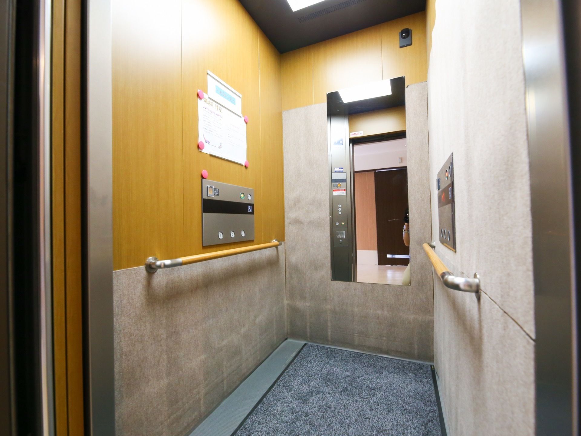 グレーと木目調を基調とした内装のエレベーター。大きな鏡、手すりが備わっている。防犯カメラが設置されている。
