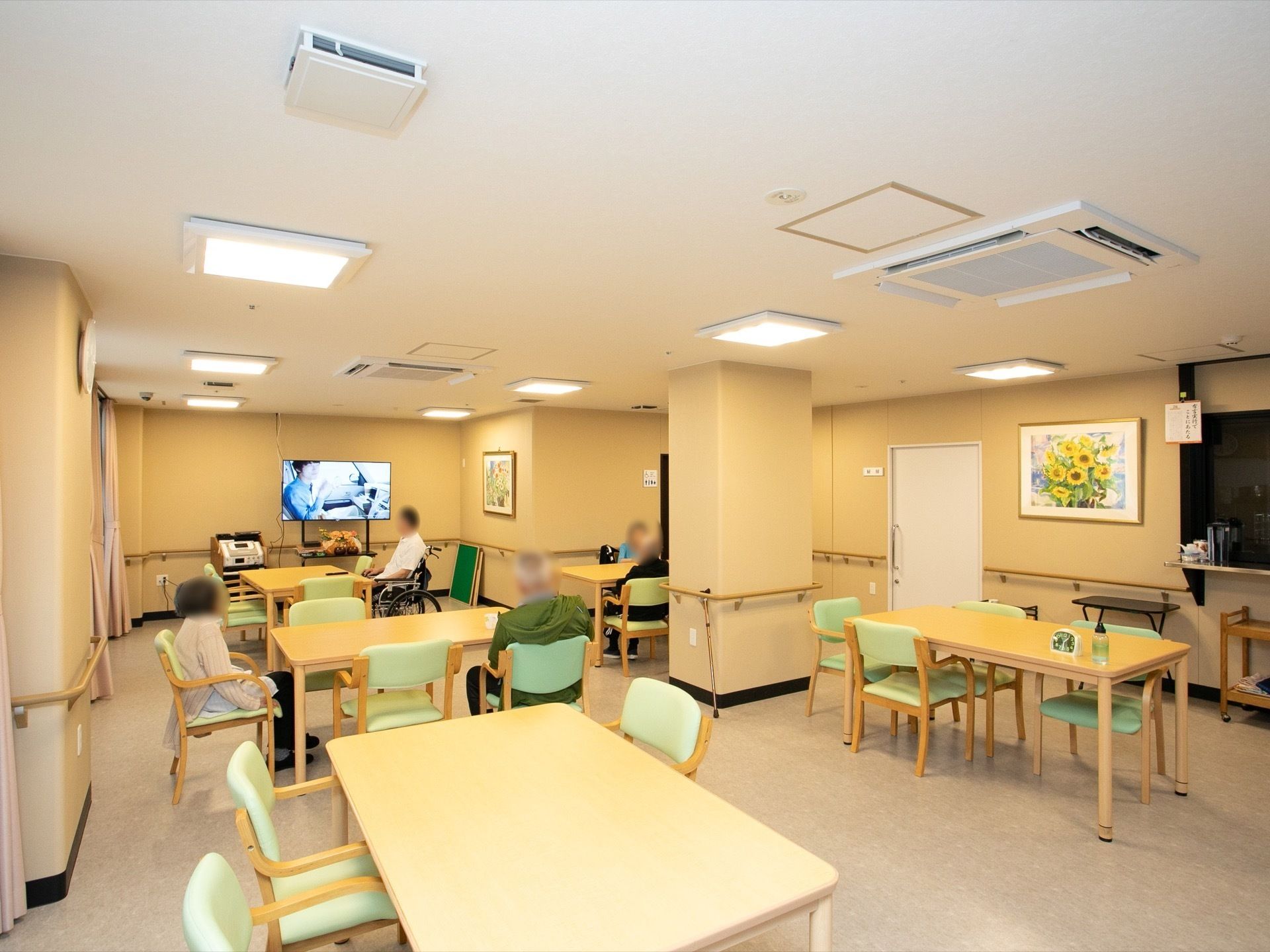 食堂にはダイニングテーブルと椅子が設置され、数人の入居者様が椅子に座っている。壁には絵が飾ってあり、奥の壁際にはテレビが置かれている。