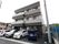 施設は3階建ての建物で、グレーやベージュの外壁になっている。建物の前には車が数台停められており、右側にはゴミ箱が置かれている。