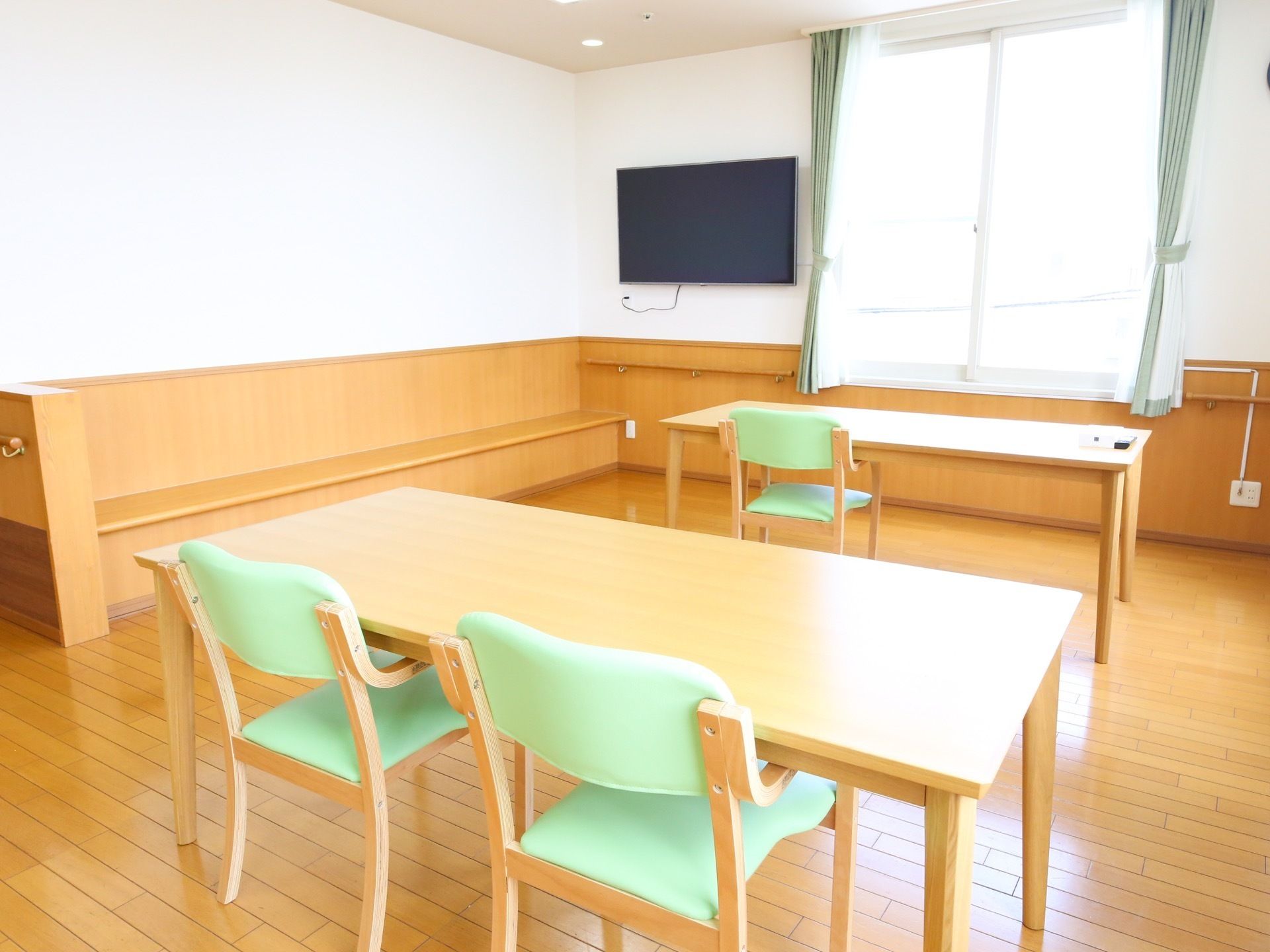 広い洋風の共有スペース。部屋の壁には大きなテレビやベンチが備えつけられている。また、部屋の中ほどに、複数のテーブルや椅子も設置されている。