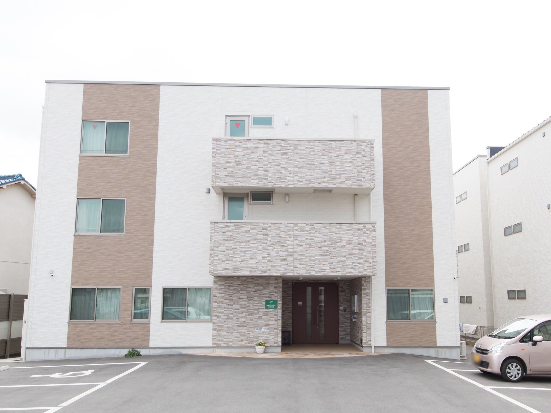 3階建ての四角い建物の前面に、広く駐車スペースが設けられている。一台ごとの区画が明確に示されており、建物の近くには優先駐車場がある。