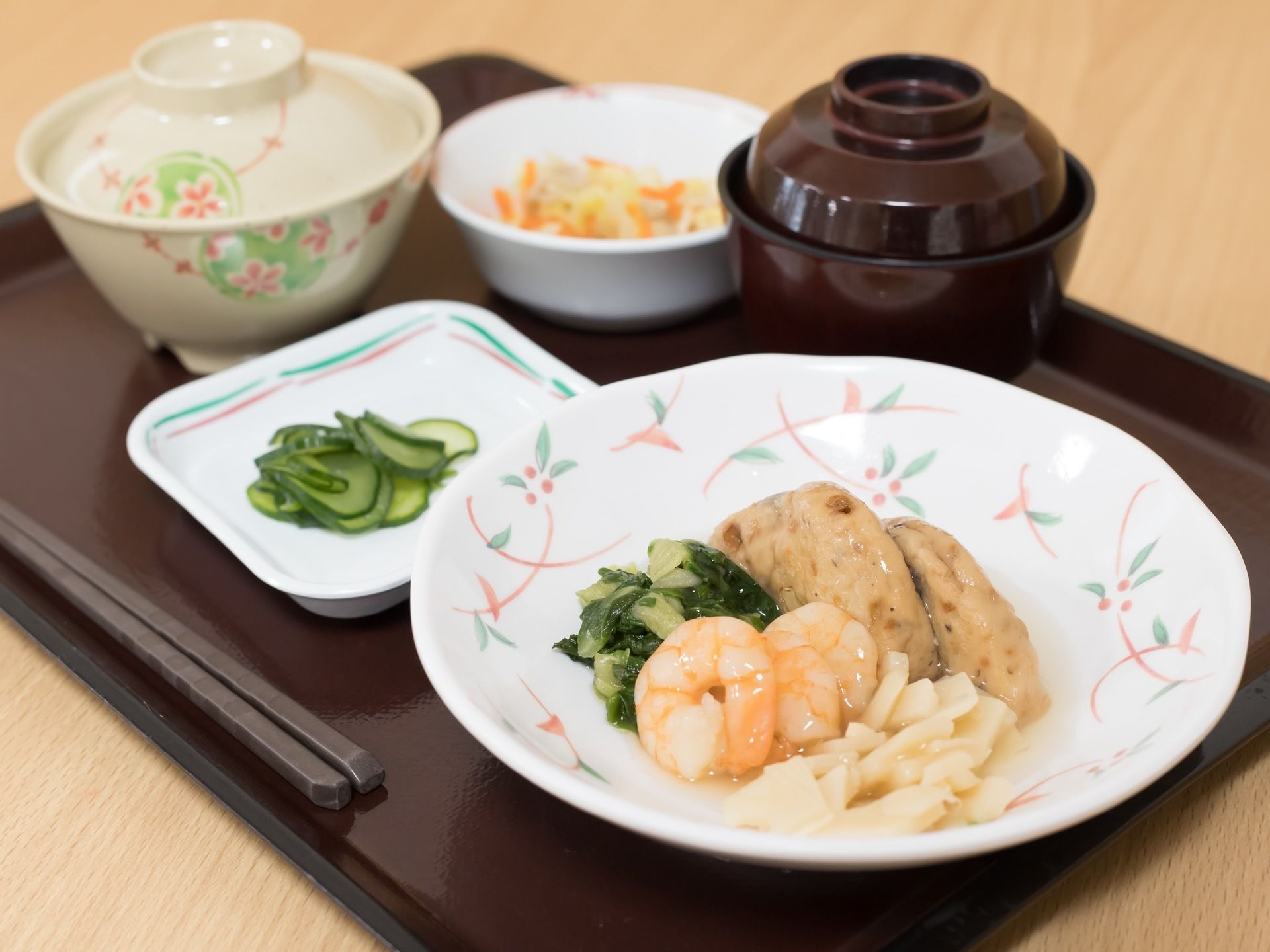 トレイの上に、蓋付きのご飯茶碗と汁椀、3種類のおかずの皿が並んでいる。煮物の小鉢、キュウリの小皿、あんかけの大皿がある。