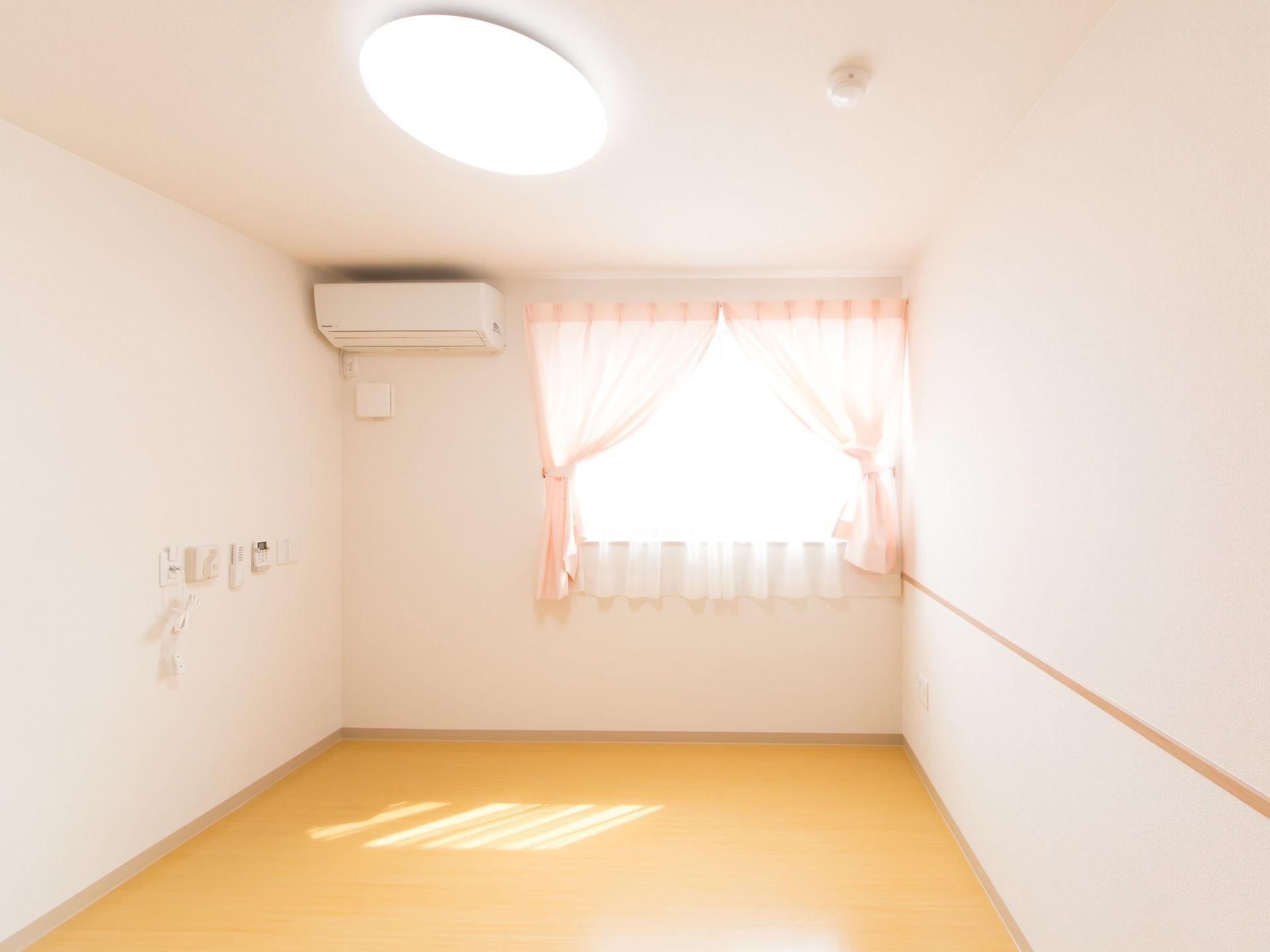 淡いピンク色のカーテンが取りつけられた窓がある。部屋の上方には、円形の大きな照明やエアコンが設置されている。