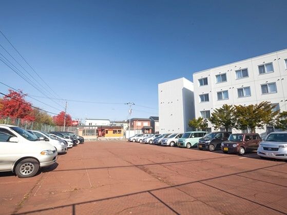 たくさんの車がきれいに並んで駐車されている駐車場。木も植えられ、周辺の住宅地も見える。右側に白い建物がある。
