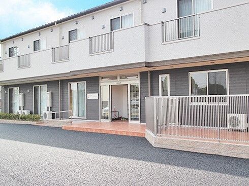 2階建ての建物は白とグレー系で建物の周りにはフェンスが立ち、玄関部分には屋根とスロープが付いている。