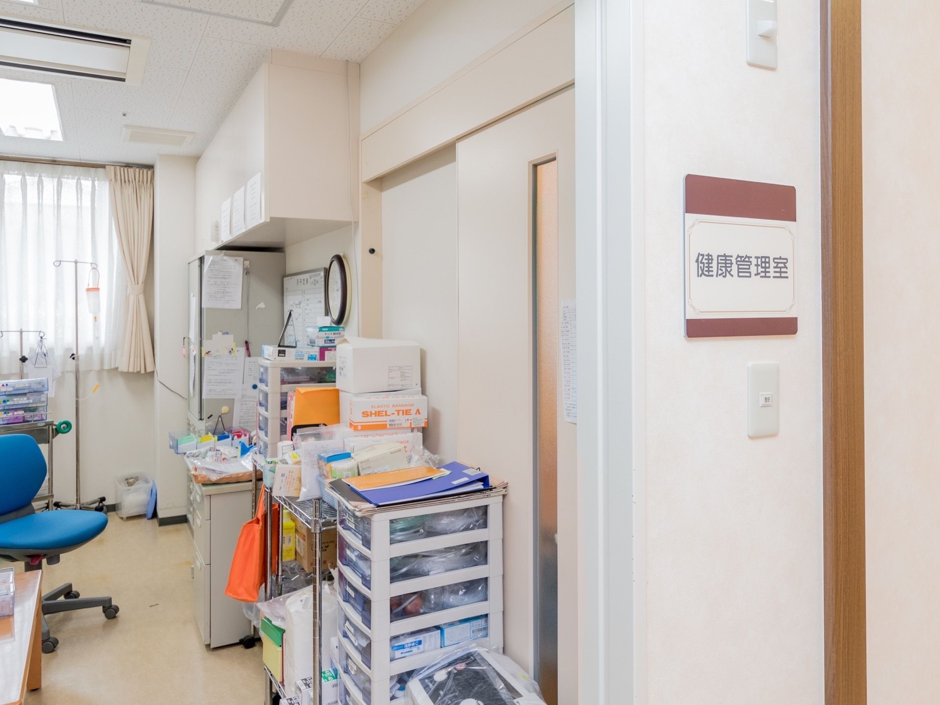 ワゴンや整理棚に収納された複数の必需品が、保管されている部屋の白い壁に設置されたルームプレート