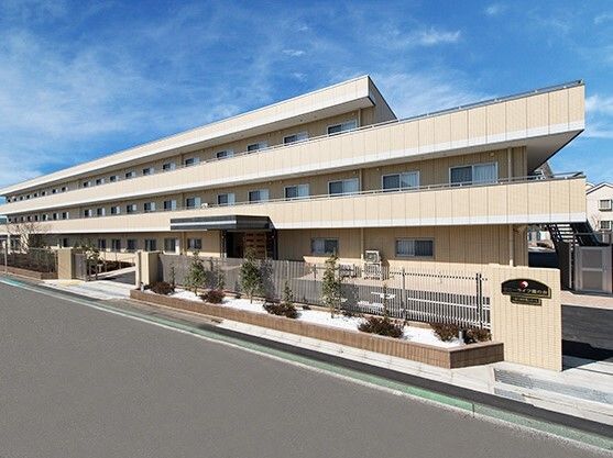 ベージュの外壁の３階建ての建物の各フロアにベランダがあり、駐車場もある。門のそばには草木が植えられている。