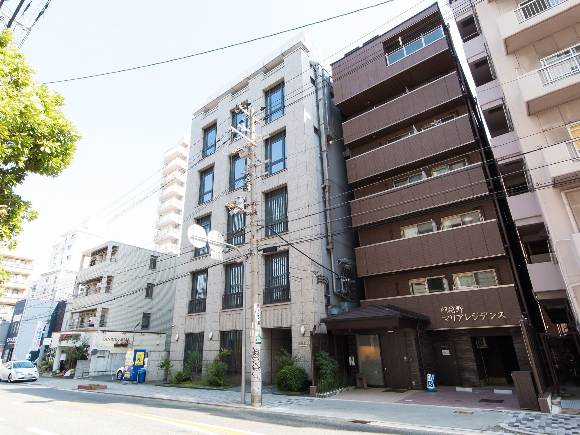 たくさんのビルが立ち並ぶ大きな通りである。歩道に電信柱があり、青い自動販売機がある。１台の車が停められている。