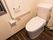 濃い色の木目の床にタンクが付いた白い洋式トイレがある。壁に手すりとナースコールとペーパーホルダーが設置されている。
