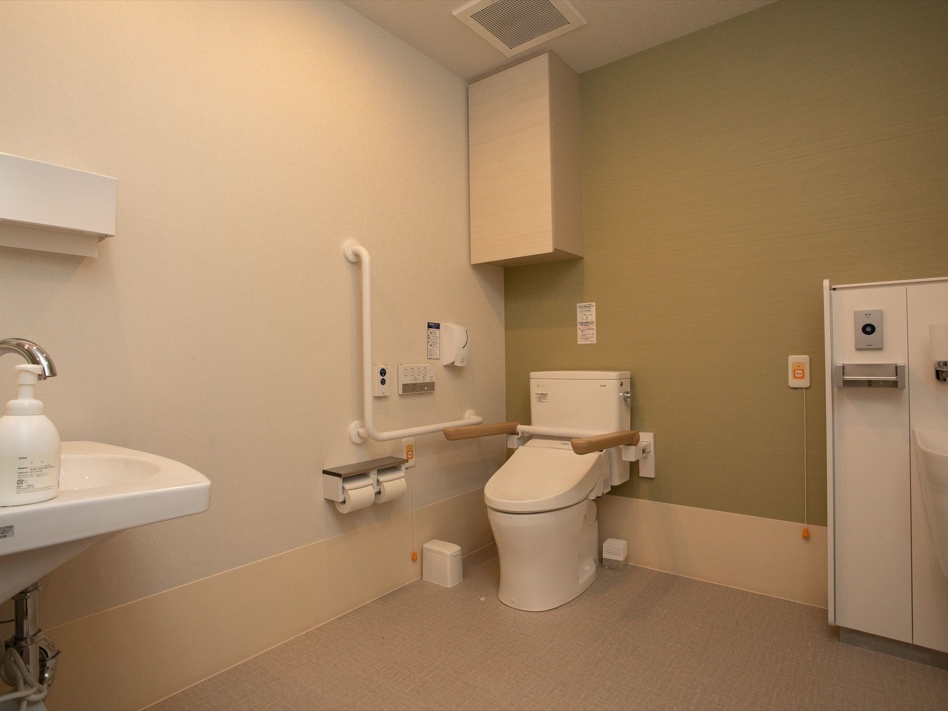 開放的な広さに設計されたトイレである。洗面台が完備されている。壁側にはＬ字形の手すりも備えつけられている。
