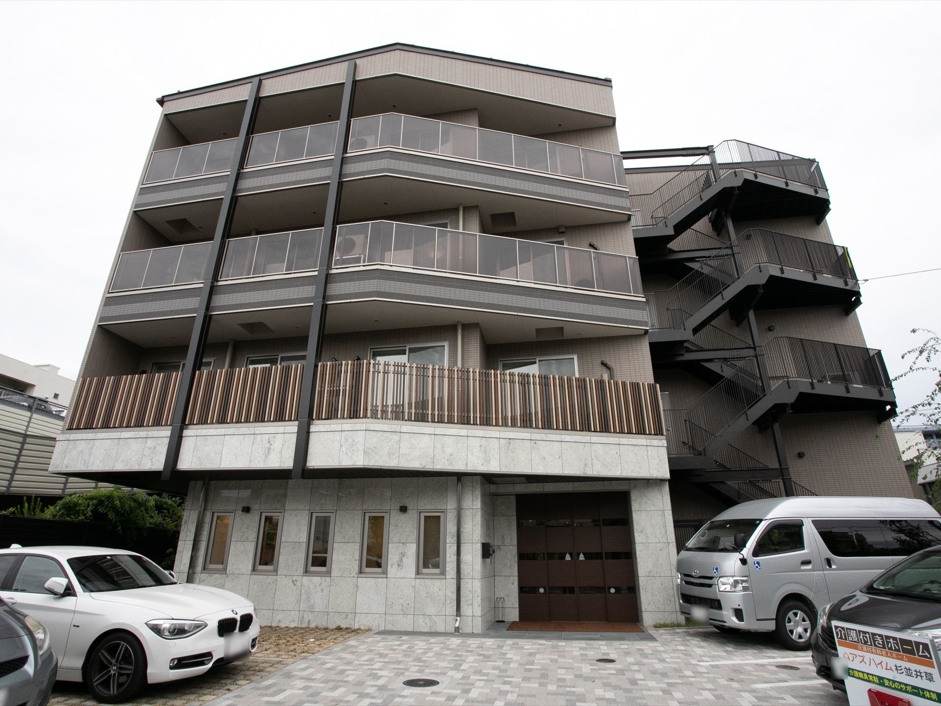 ブラウンを基調とした4階建ての建物は玄関周りに駐車場や木が植えられたスペースがあり、すっきりとした印象。