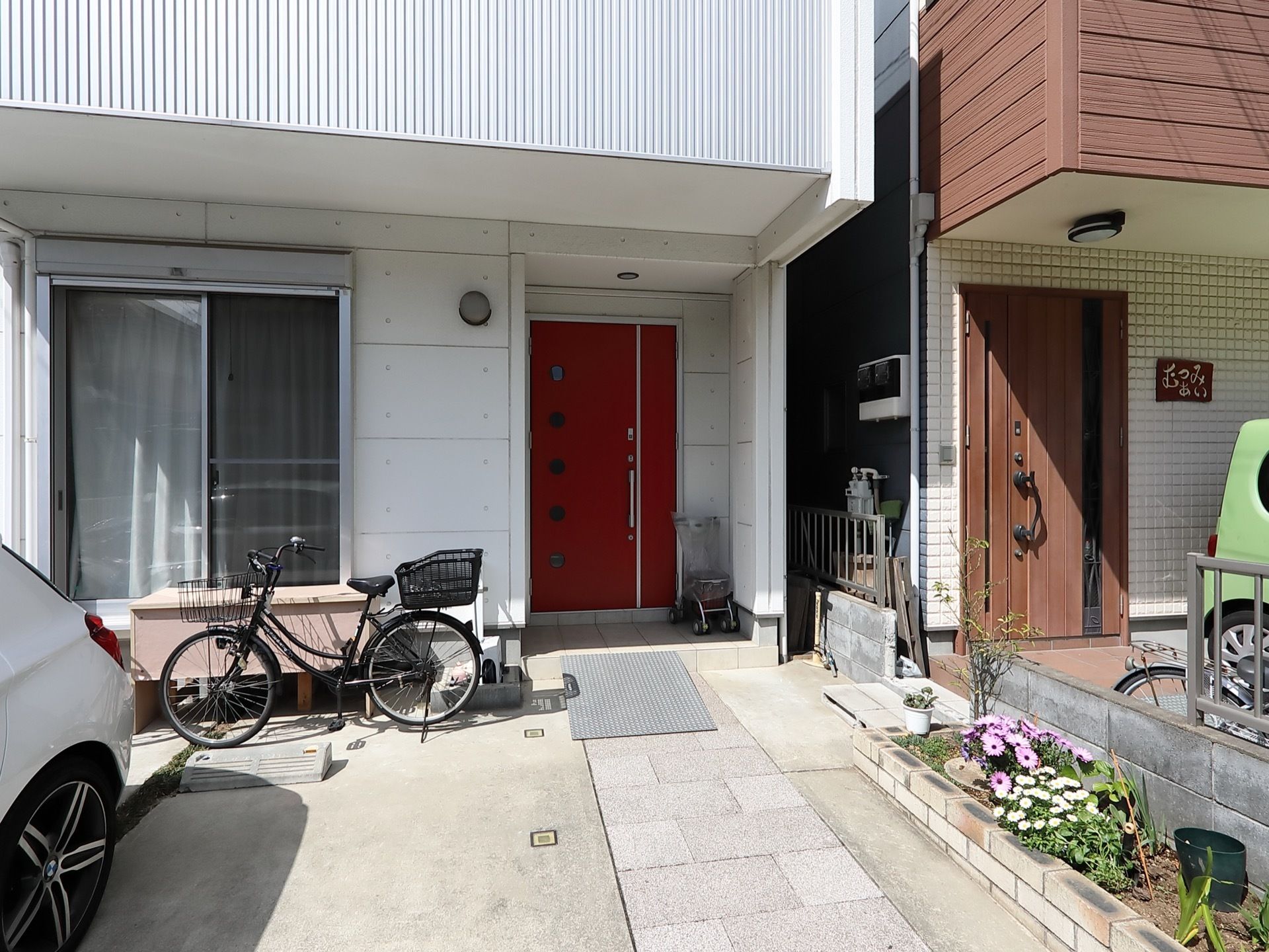 白を基調とした一軒家の建物になっている。玄関扉は鮮やかな赤色である。自転車が1台置かれており、車が停まっている。