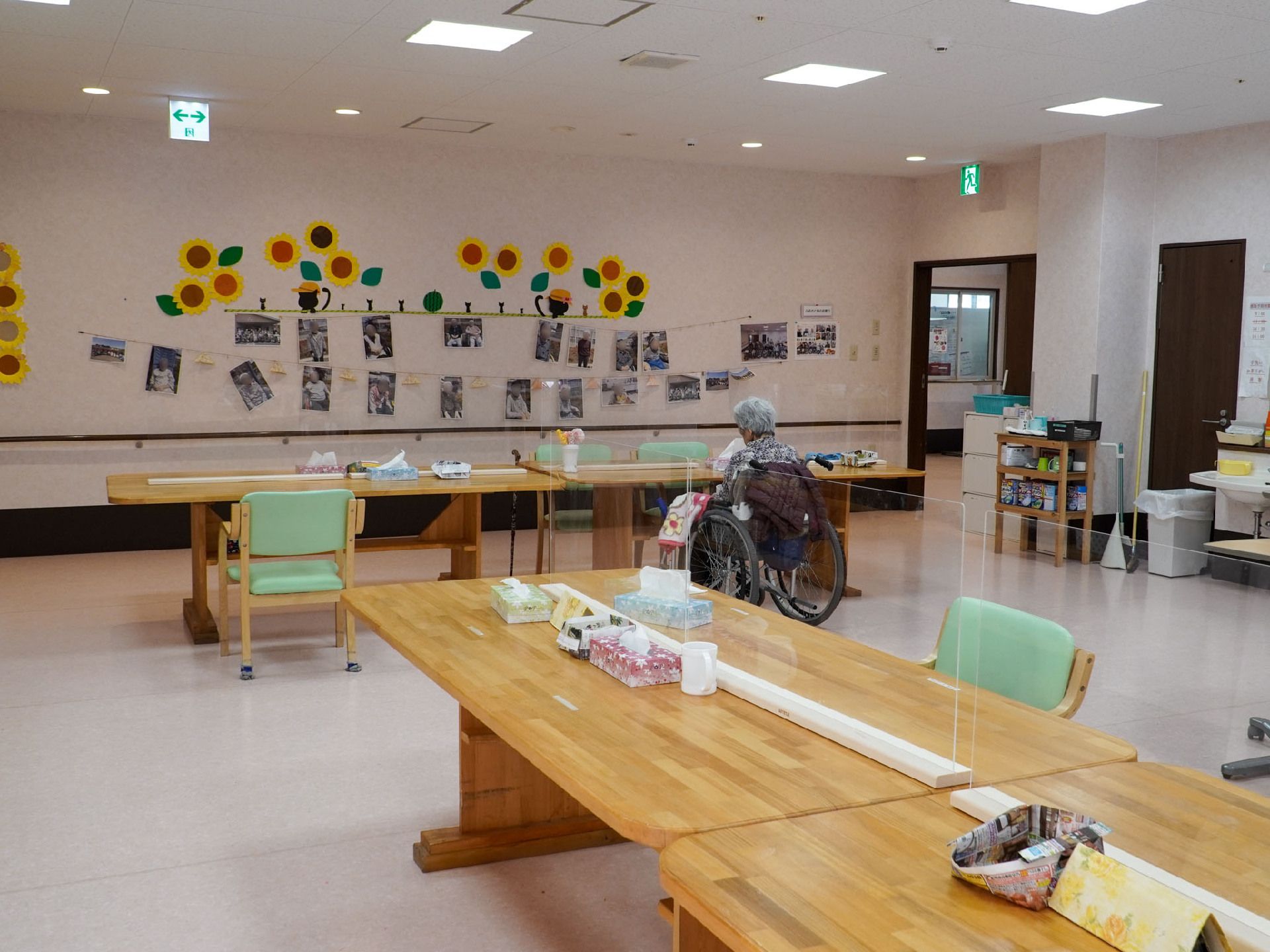 仕切りのある長テーブルが置かれた部屋の壁に貼られた写真を見ている車いすの入居者様