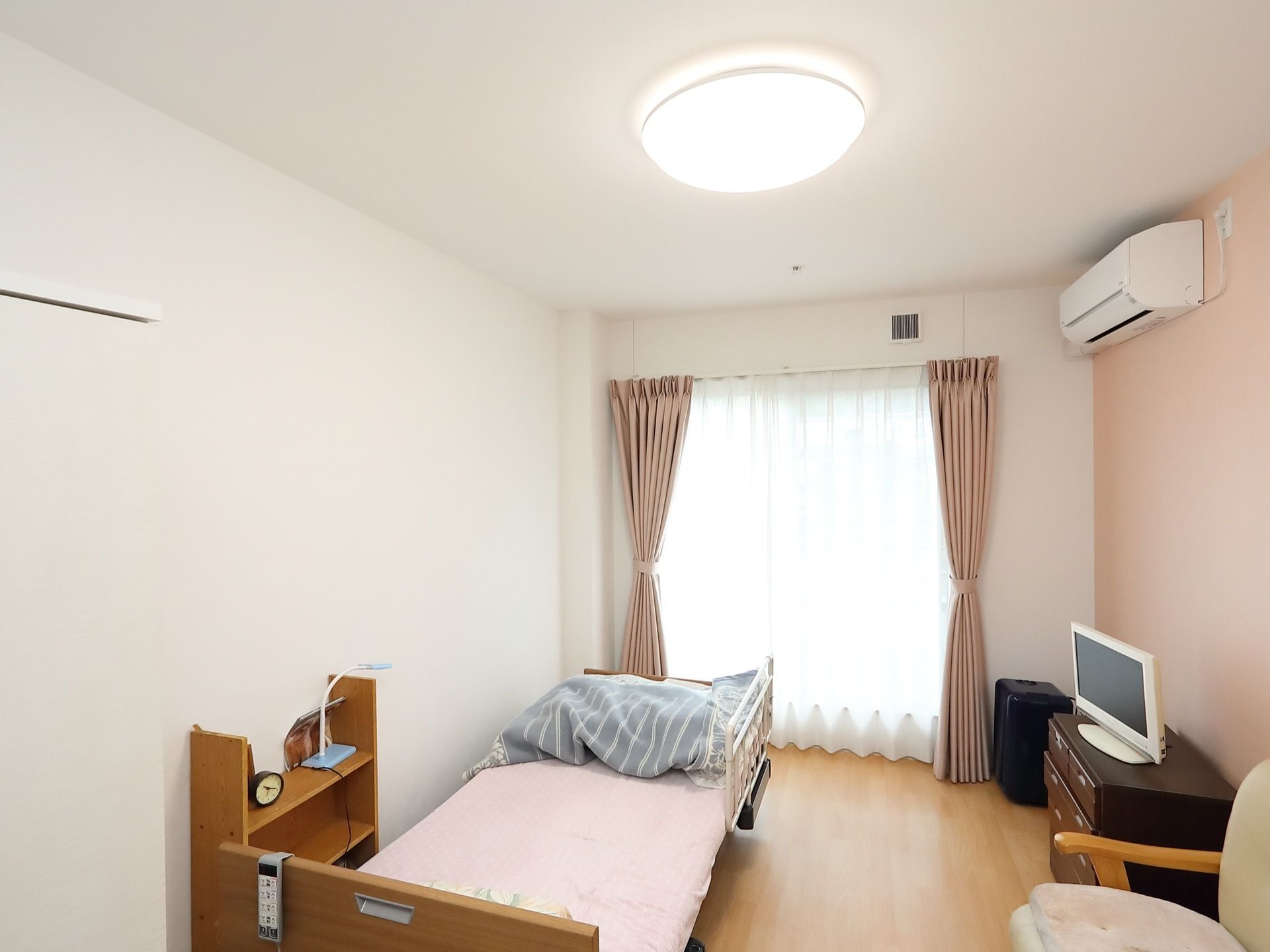 壁紙とカーテン、ベッドのシーツの色が調和していて美しい空間。木製の収納家具の上に液晶モニターが置かれている。