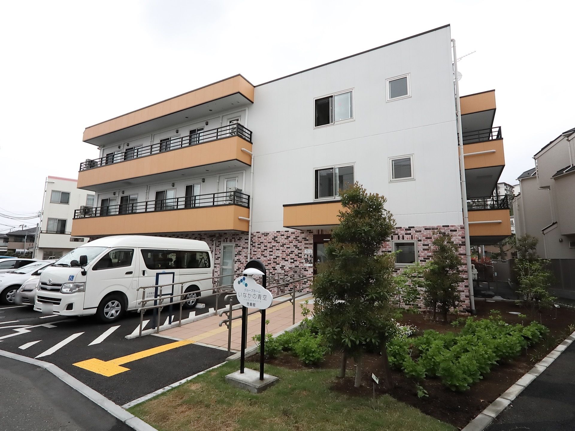 ３階建ての施設の前面がアスファルトで舗装されており、複数の自動車が駐車している。玄関から続くスロープの脇に、草花や庭木が植えてある。