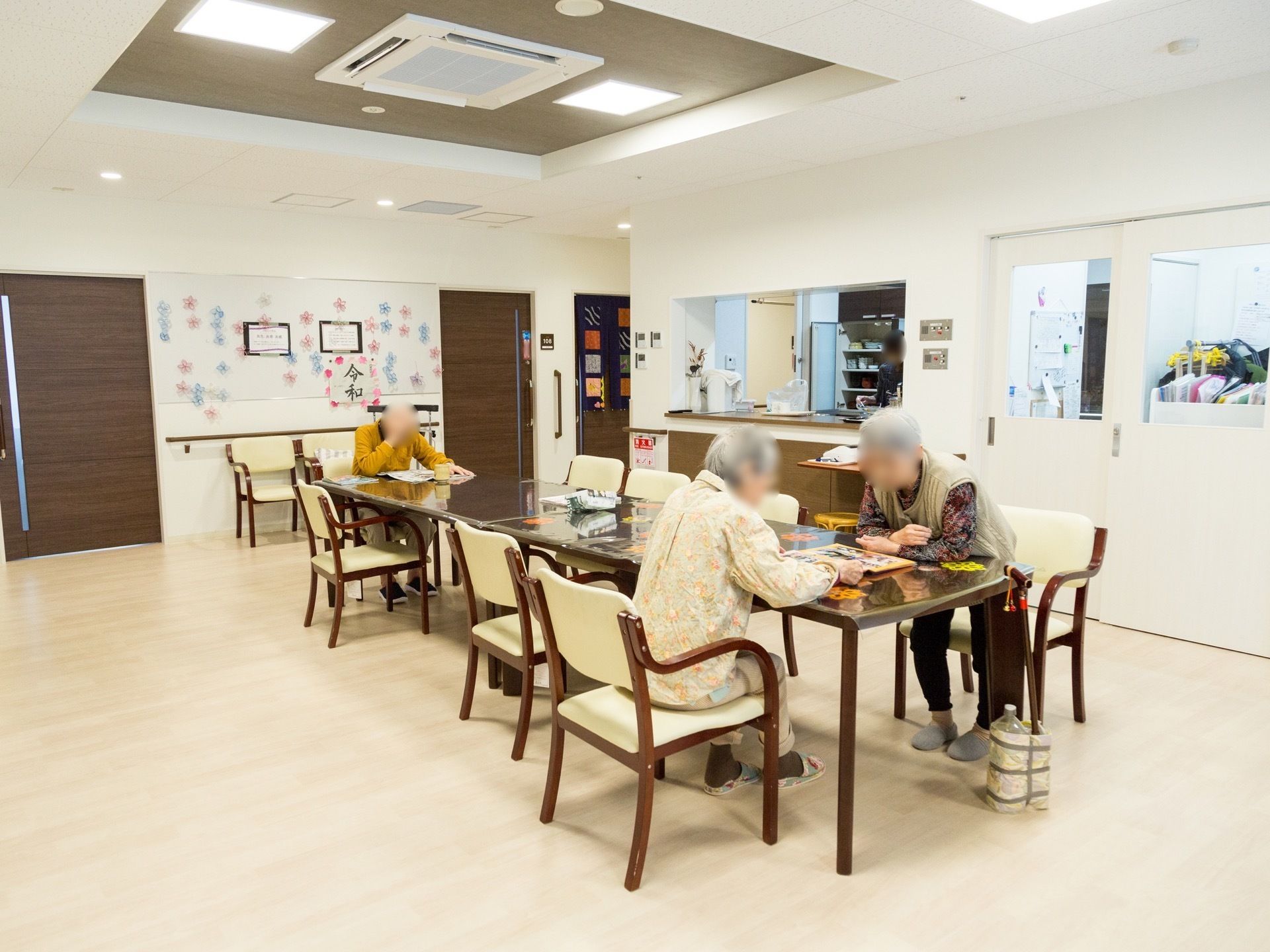 ４人がけテーブルを２セット並べ、８名から１０名までの入居者様が席につけるようにしている。スタッフルームやキッチンには、窓が付いている。