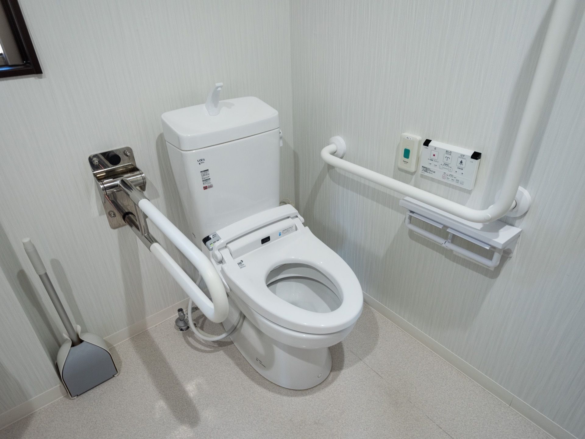 温水洗浄便座が完備されたトイレである。壁には、操作パネルや緊急呼び出しブザー、L字型手すりが設置されている。