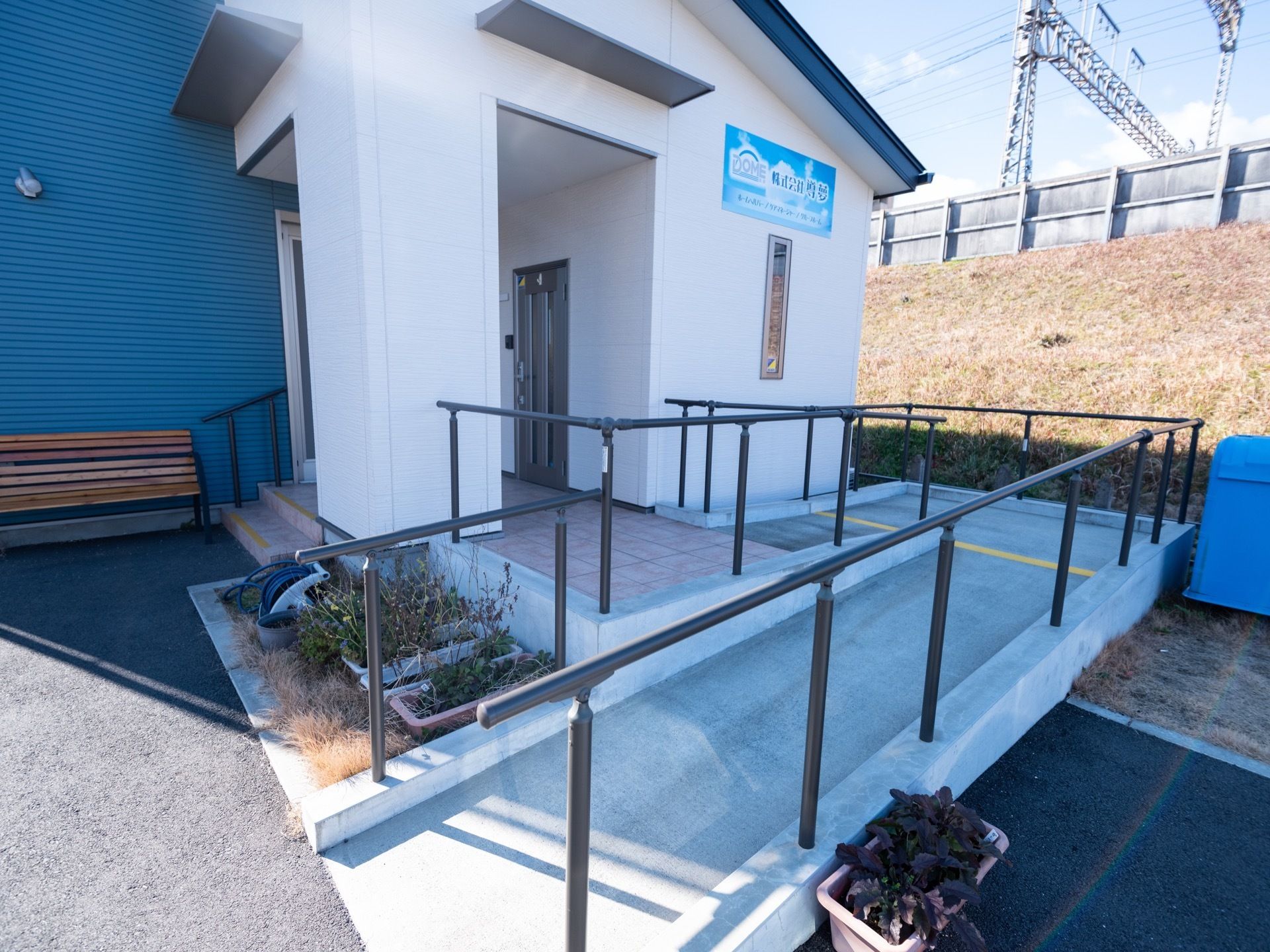 施設入口には、手すりが備え付けられたスロープが完備されている。奥には、木製のベンチが置かれているのが見える。