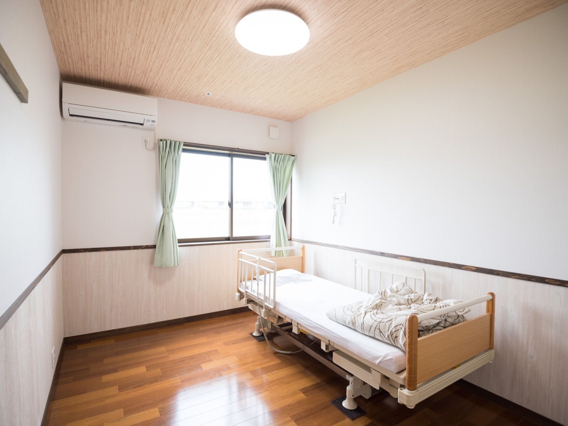 大きな窓があり、エアコン、手すりつき介護ベッドがある居室である。床と天井に木を使い、温かみが感じられる。