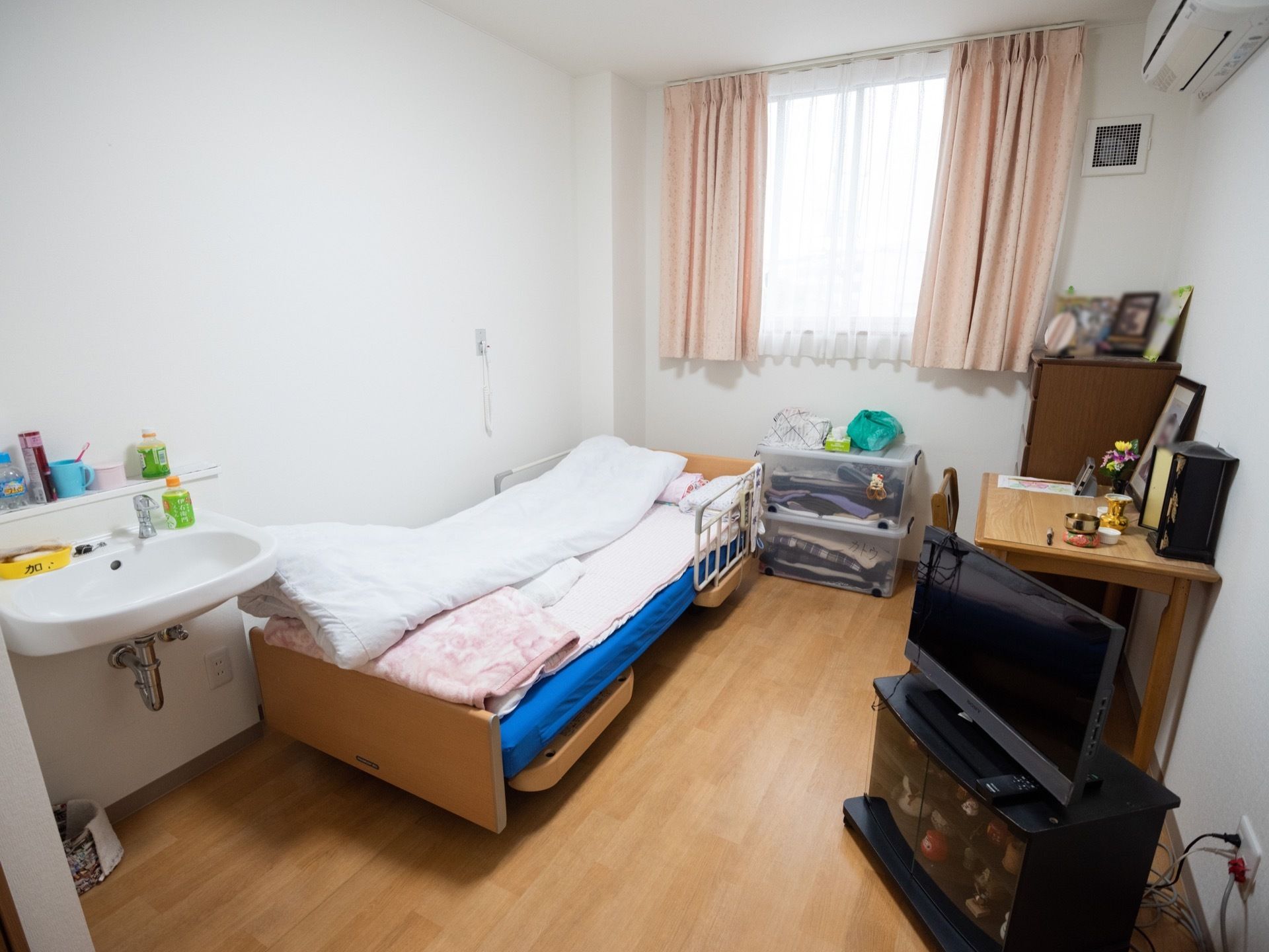 洋風の部屋の中に、手洗い場やエアコンが備え付けられ、介護ベッド、テレビ、収納、デスク、椅子が置いてある。