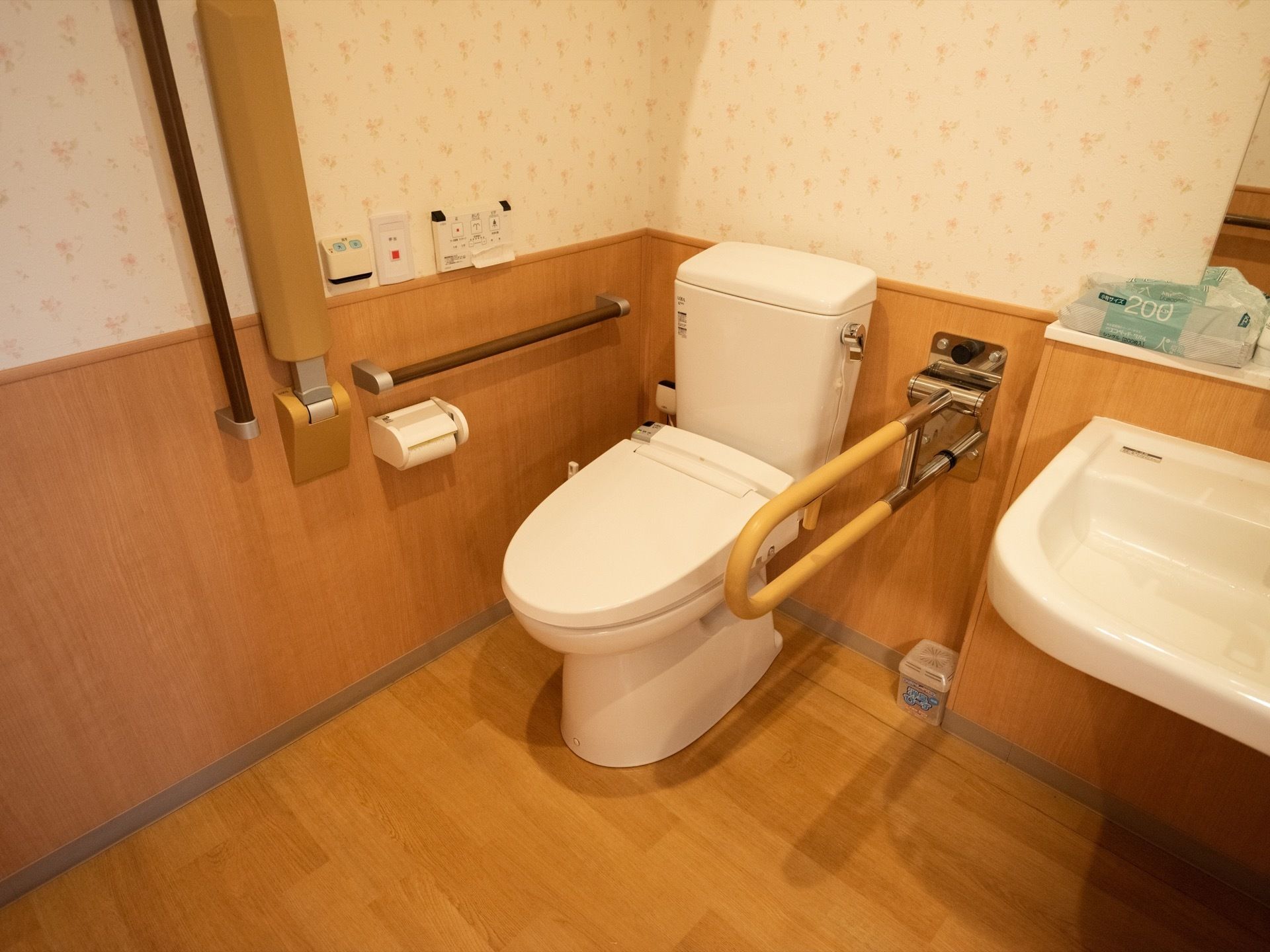 洋式のトイレと洗面所のあるバリアフリーのトイレの個室。緊急呼出しボタンと可動式の手すり、温水洗浄便座のリモコンなどが備えられている。