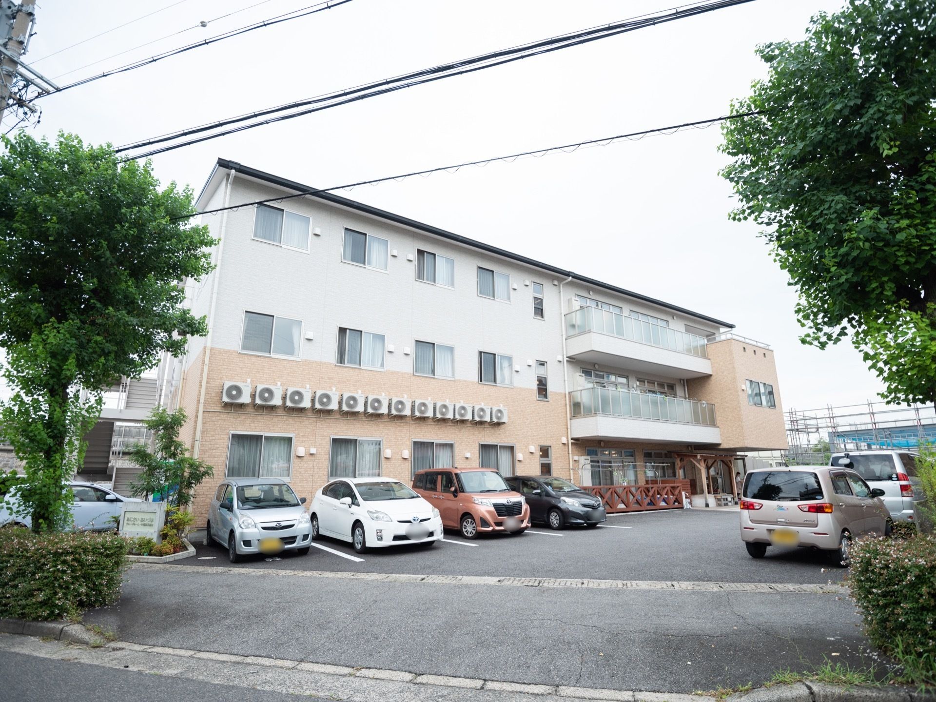 街路樹のある道路沿いに建つ、３階建てのホーム。ホームの敷地内には駐車場やホームの名称の書かれたコンクリートの看板がある。