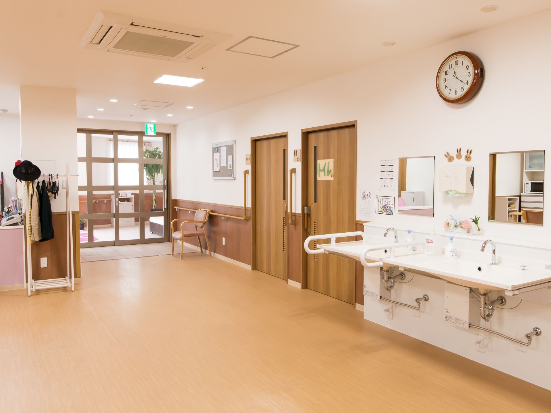 トイレのドアのすぐ横に洗面台が２台並んである。壁には鏡と時計が掛かっている。台の周囲には手すりがある。