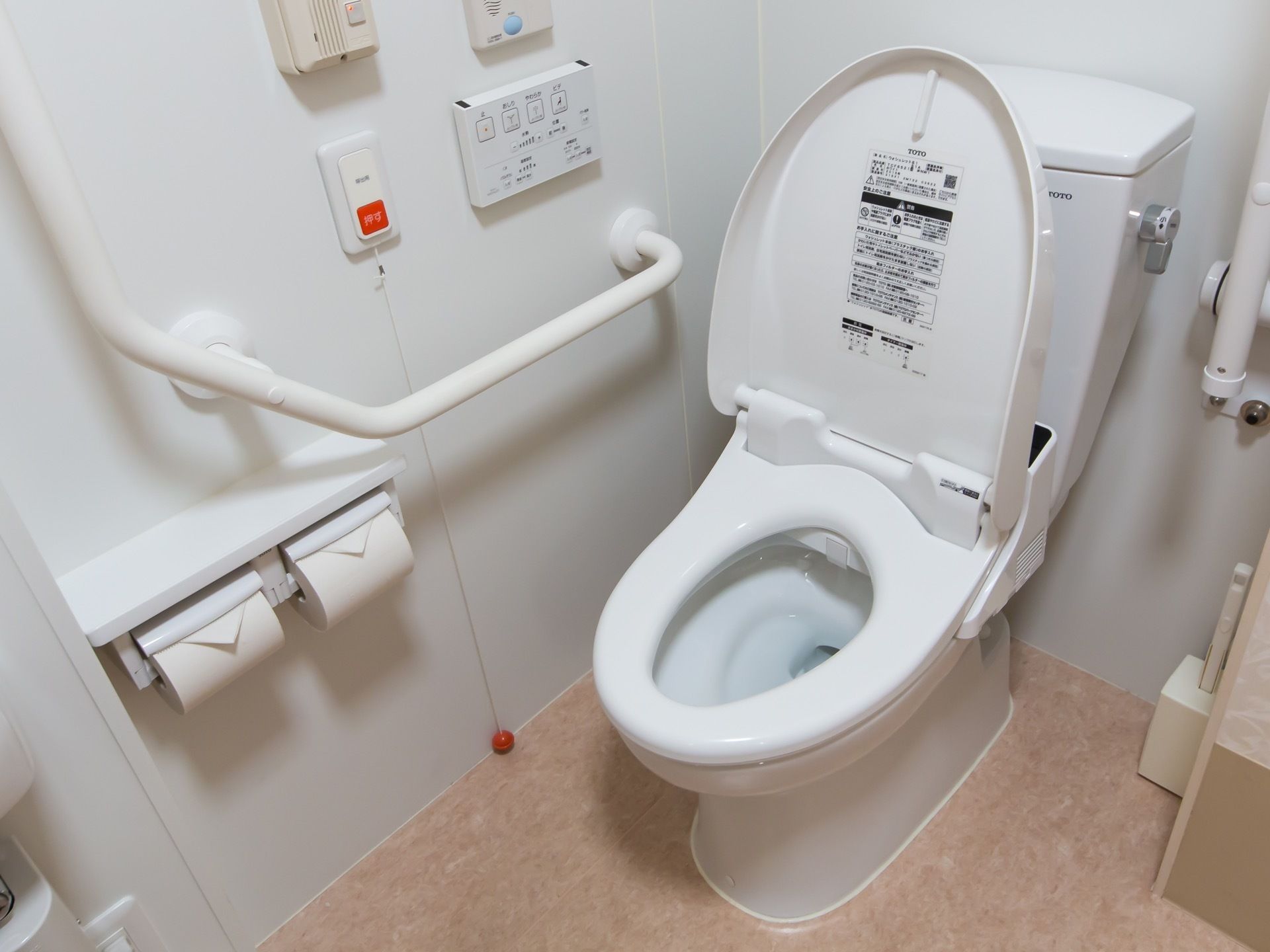 白い壁とベージュの床のトイレがある。壁には大きな手すりがある。便器には蓋があり、温水洗浄便座が付いている。