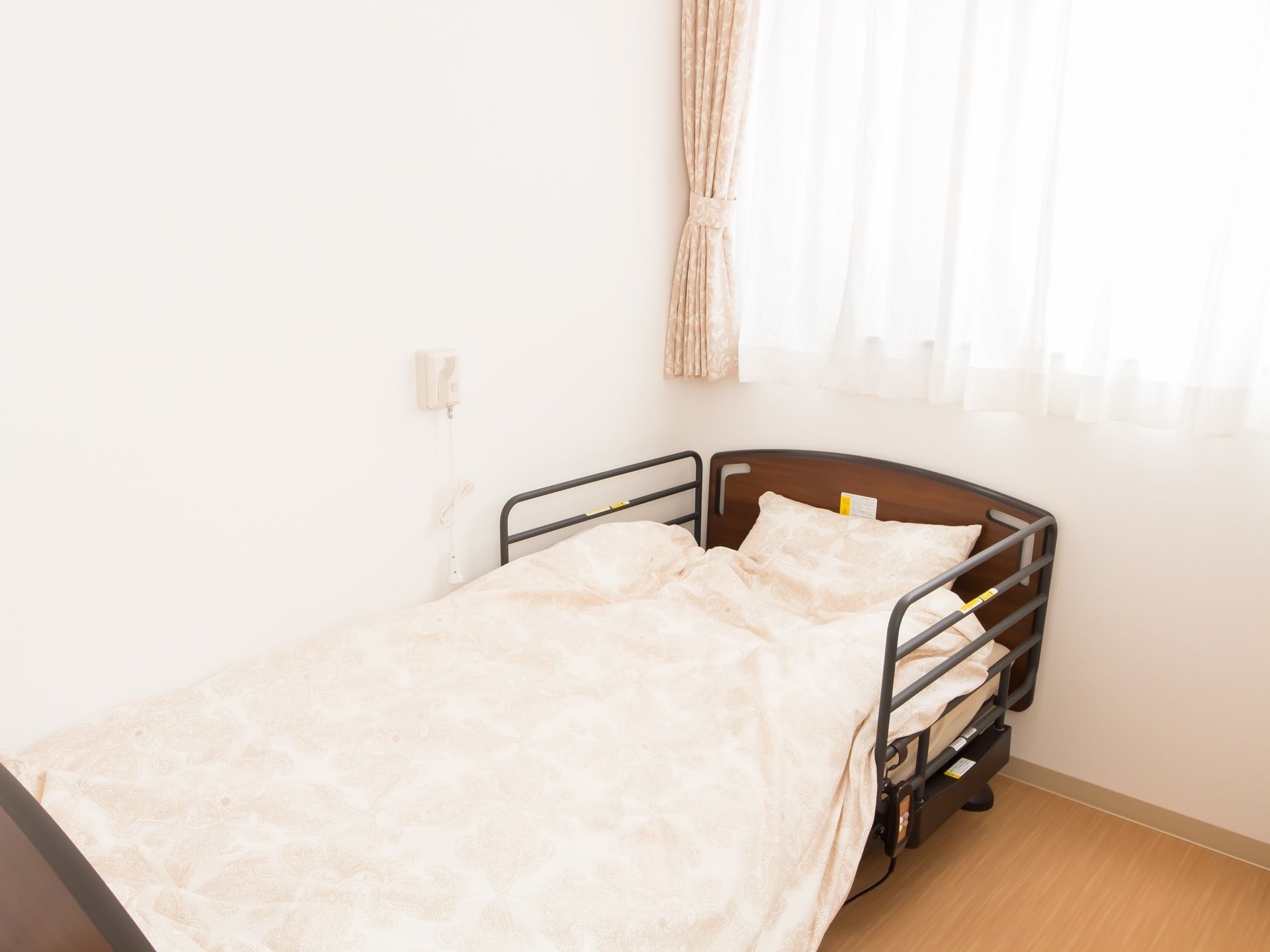 白い壁と大きな窓のある居室。窓にはカーテンが掛かっており、側には柵の付いた介護用ベッドがある。