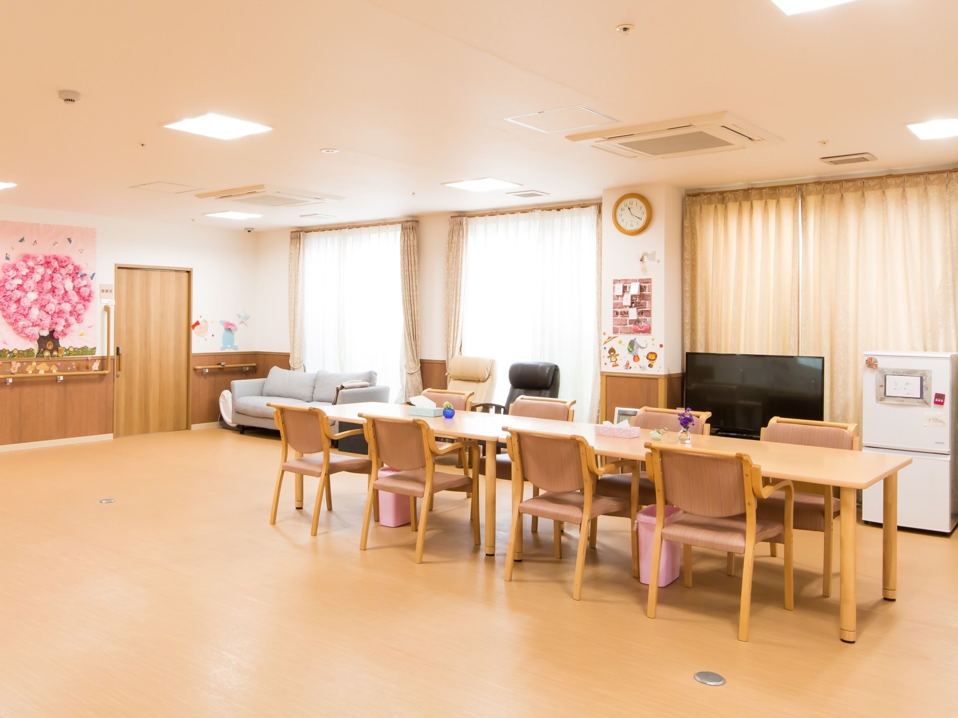 壁一面が窓になっている広い食堂がある。テーブルと椅子が一列に並んでいる。窓際にはソファとテレビが置かれ、壁には手作りの飾りつけがされている。