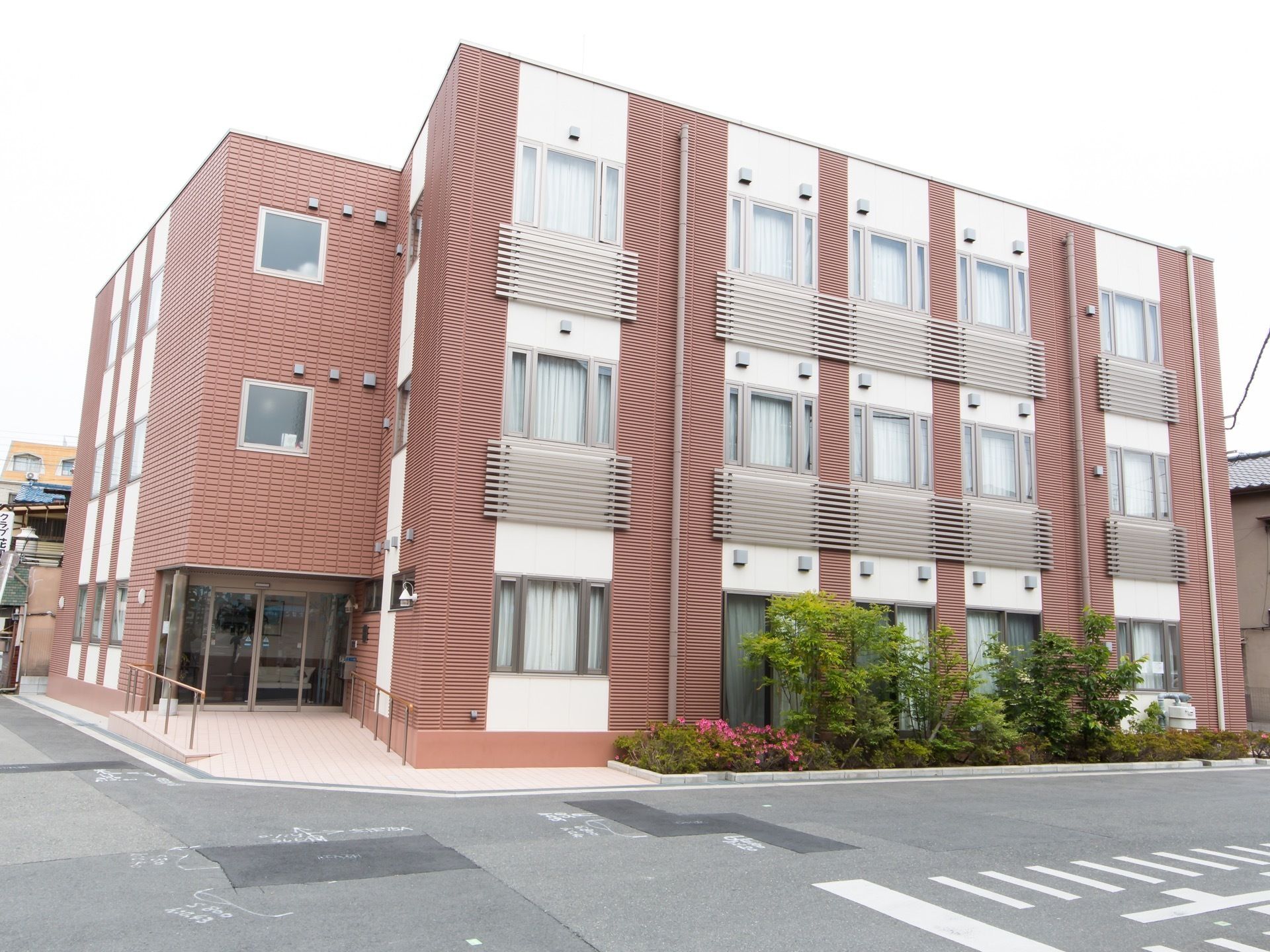 広い道路の角地に面した場所に建っている３階建ての建物。赤茶色のタイルと白いポイントの付いた壁になっている。緑の繁っている植え込みがある。