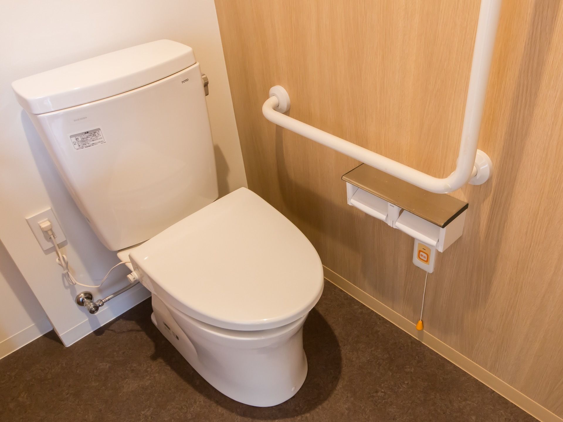 白い水洗トイレが設置されており、壁にはＬ字型の手すりやトイレットペーパーホルダー、呼び出しボタンが取りつけられている。