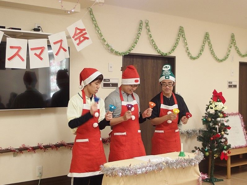 男性スタッフがおそろいの赤いエプロンとサンタ帽をかぶり、クリスマスツリーのそばでハンドベルを演奏している。