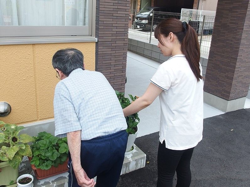髪の長い女性スタッフに付き添われて、敷地内に置かれたプランターの植物を見ている高齢男性。