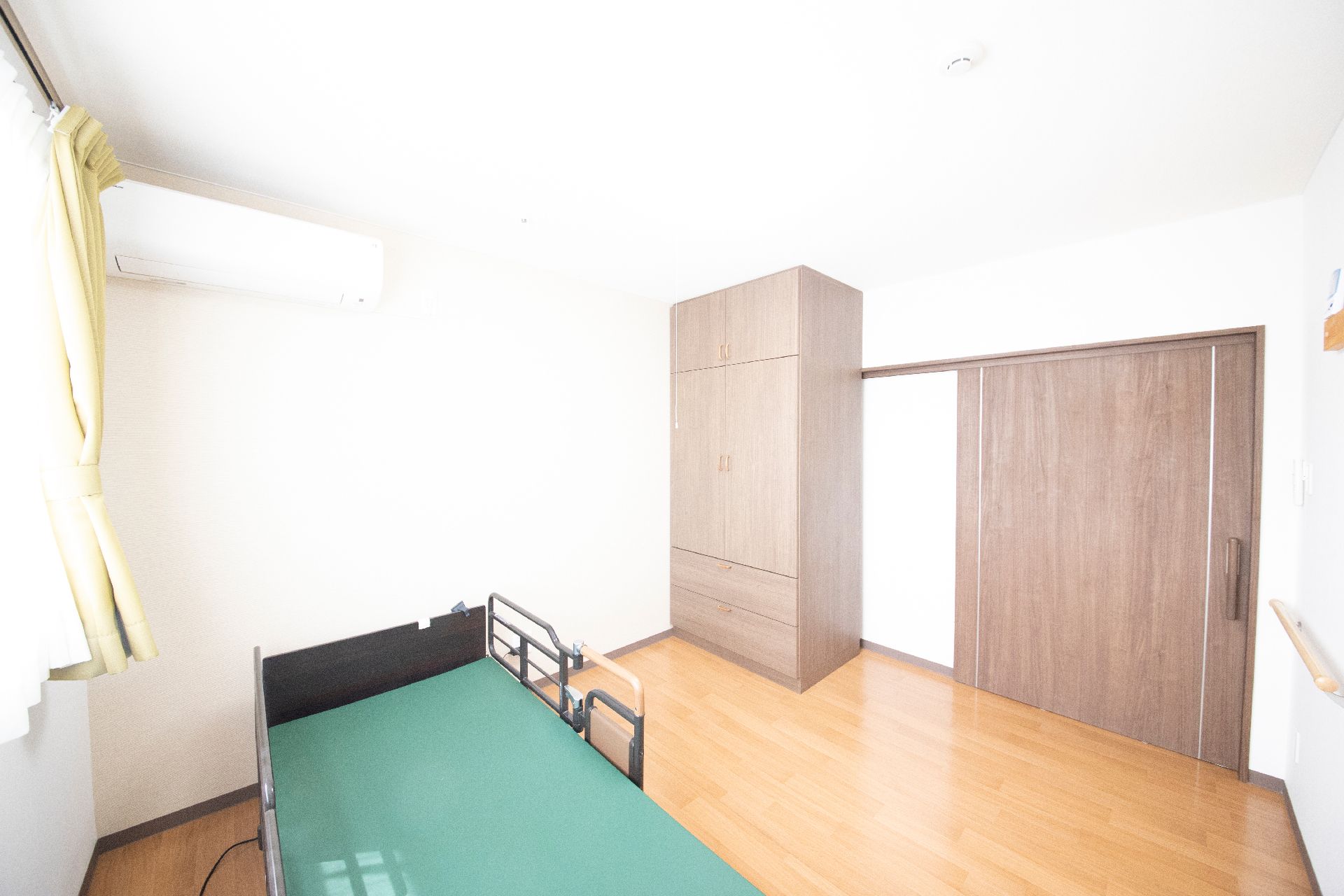 緑の多い屋外でエプロンを付けた女性スタッフに付き添われて外気浴をしている車いすの高齢女性。