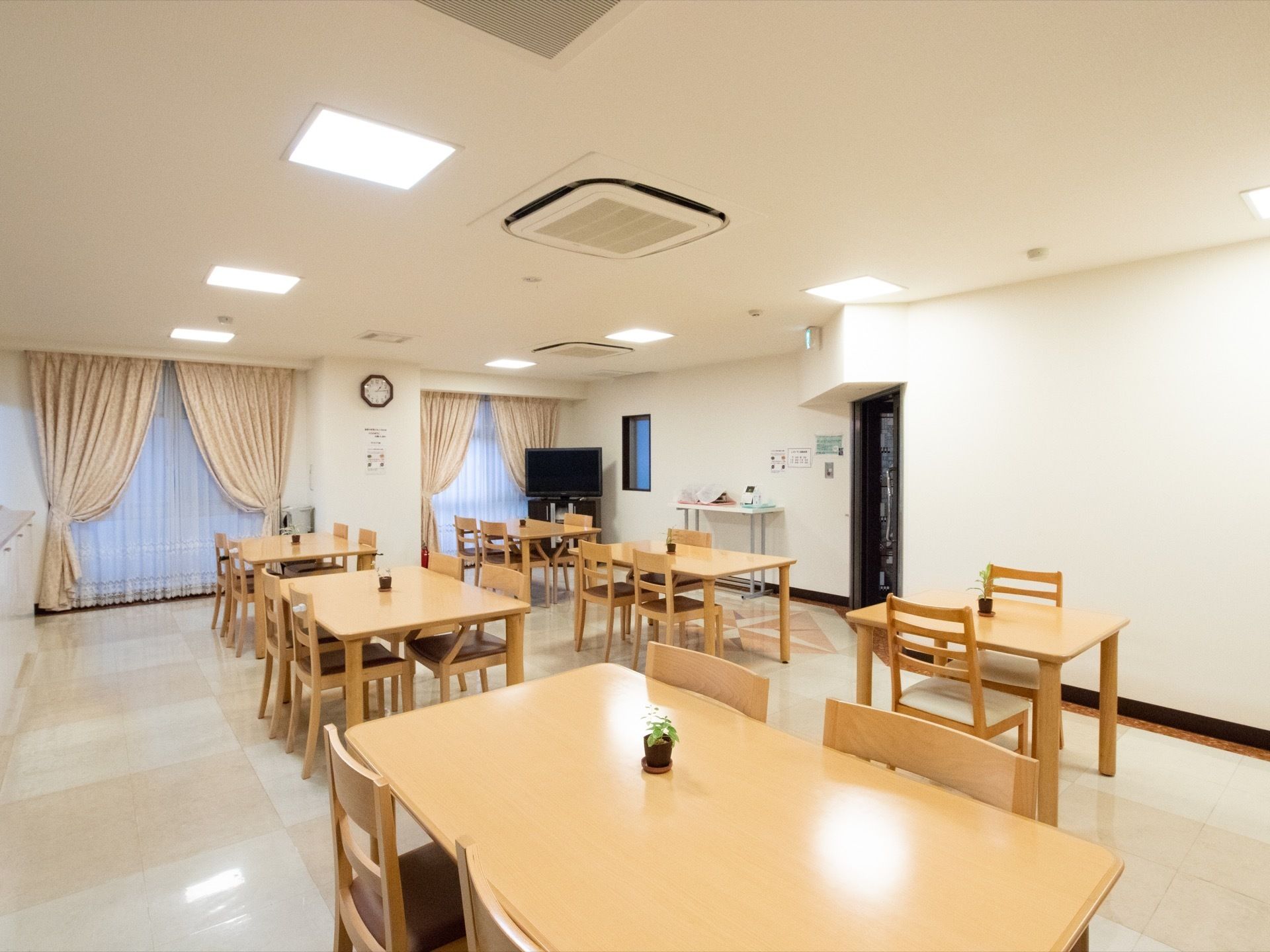 食堂の隅にはテレビが設置され、白い壁には時計や掲示物が掛けられている。テーブルはナチュラルな木目調である。