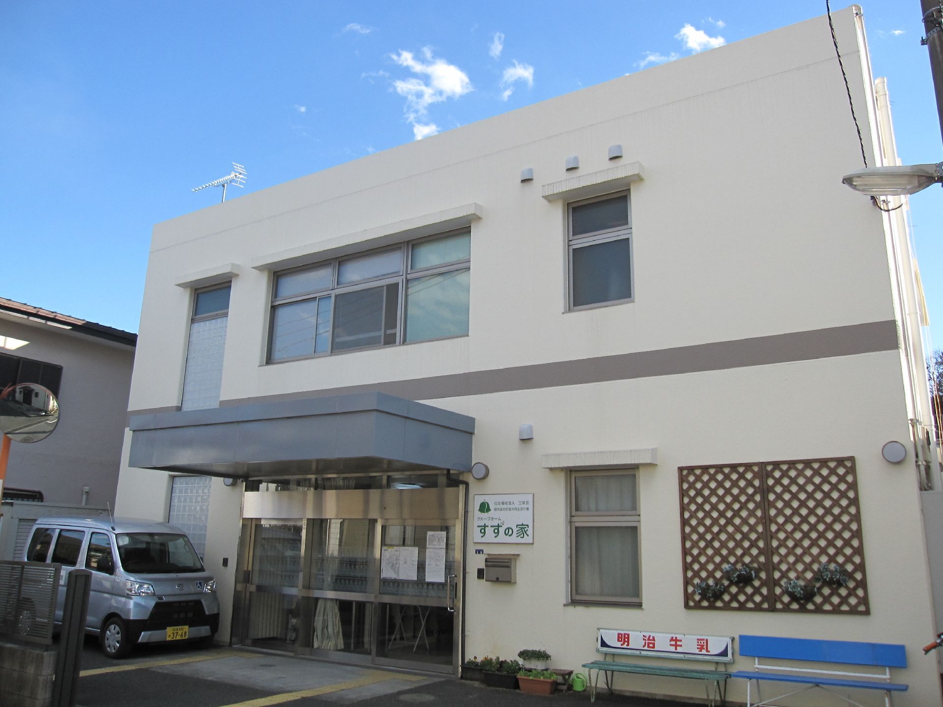 原っぱのような場所を散歩しているブラウンのジャケットを着た高齢女性と付き添いのエプロンを付けた若い女性スタッフ。