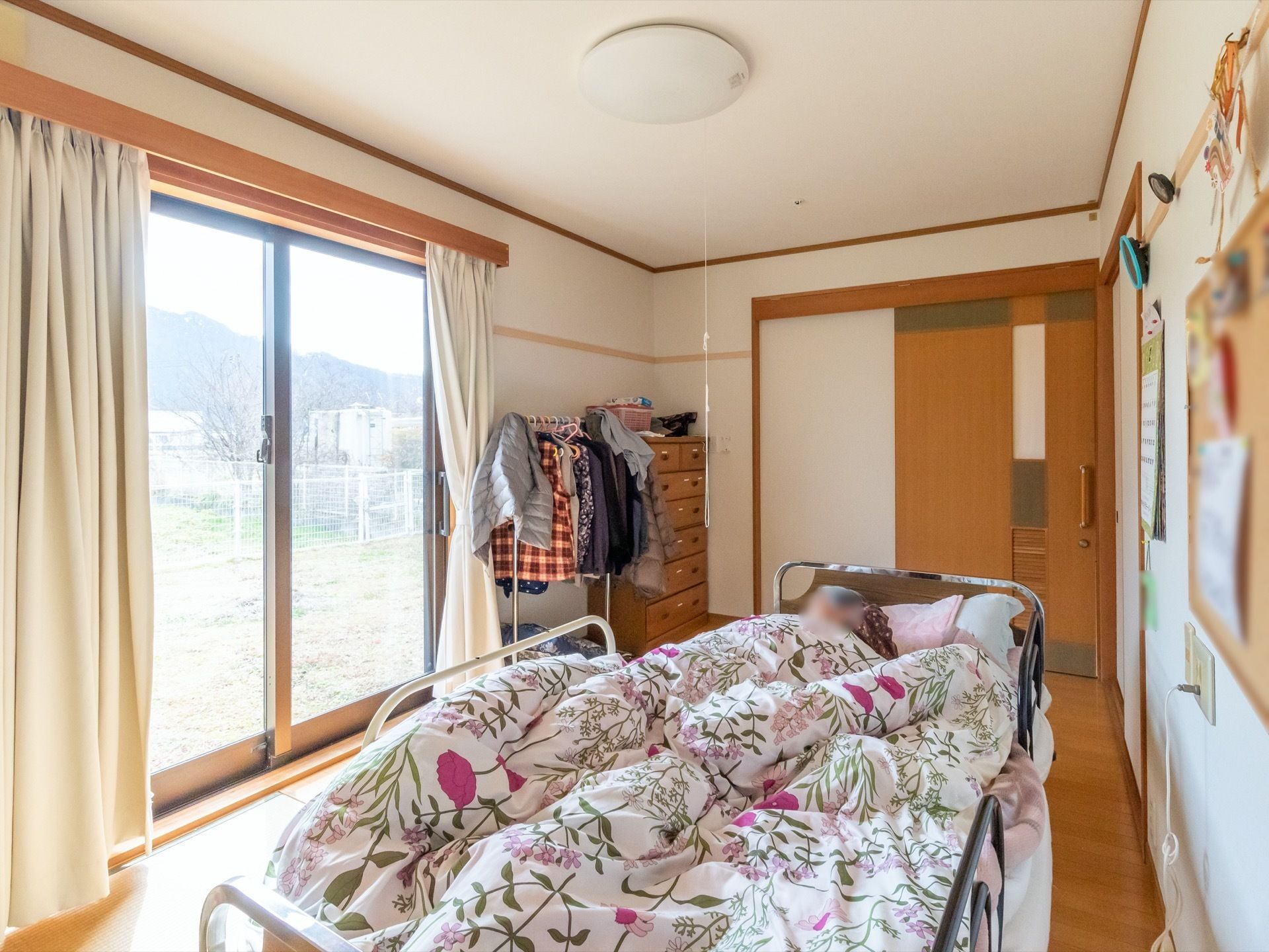 部屋の中央にベッドが１台置かれており、窓にはカーテンが取り付けられている。窓からは、外の景色が見える。