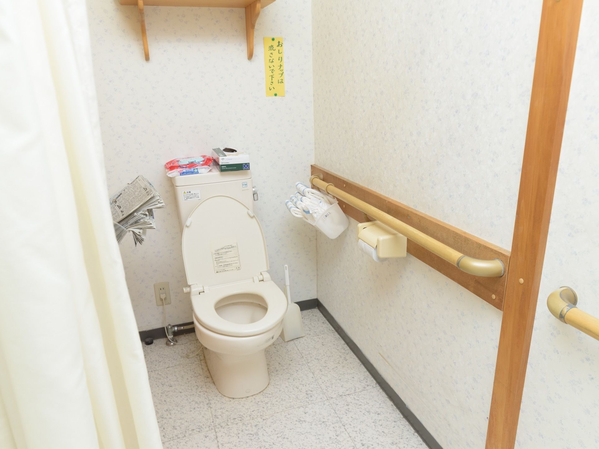 ホーム内にあるトイレの個室。壁には手すりが備え付けられているほか、各種の介護用品や、新聞紙の束が入ったカゴなども吊り下げられている。