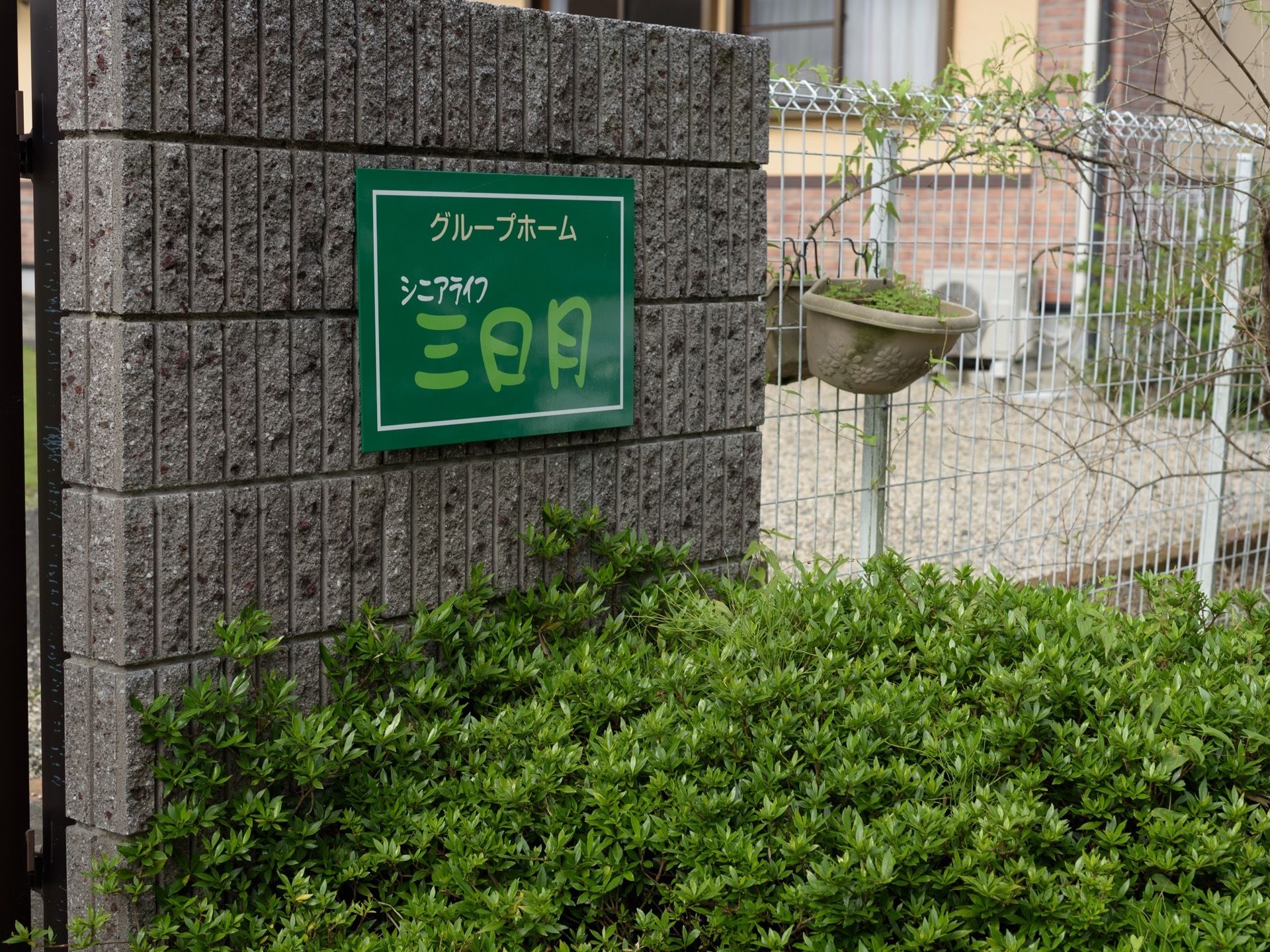 ホームの敷地を囲う白い金属製のフェンスがあり、その出入口の門扉の横に、ホームの名称が表示された看板が設置されている。